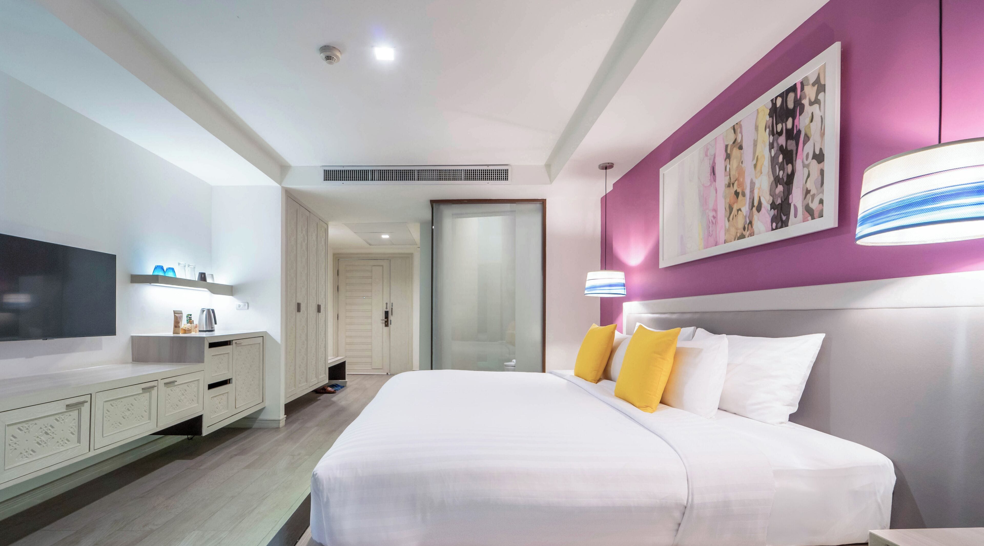 A modern hotel room with a large bed, a flat-screen TV, a bedside table with a lamp, and a large window.
