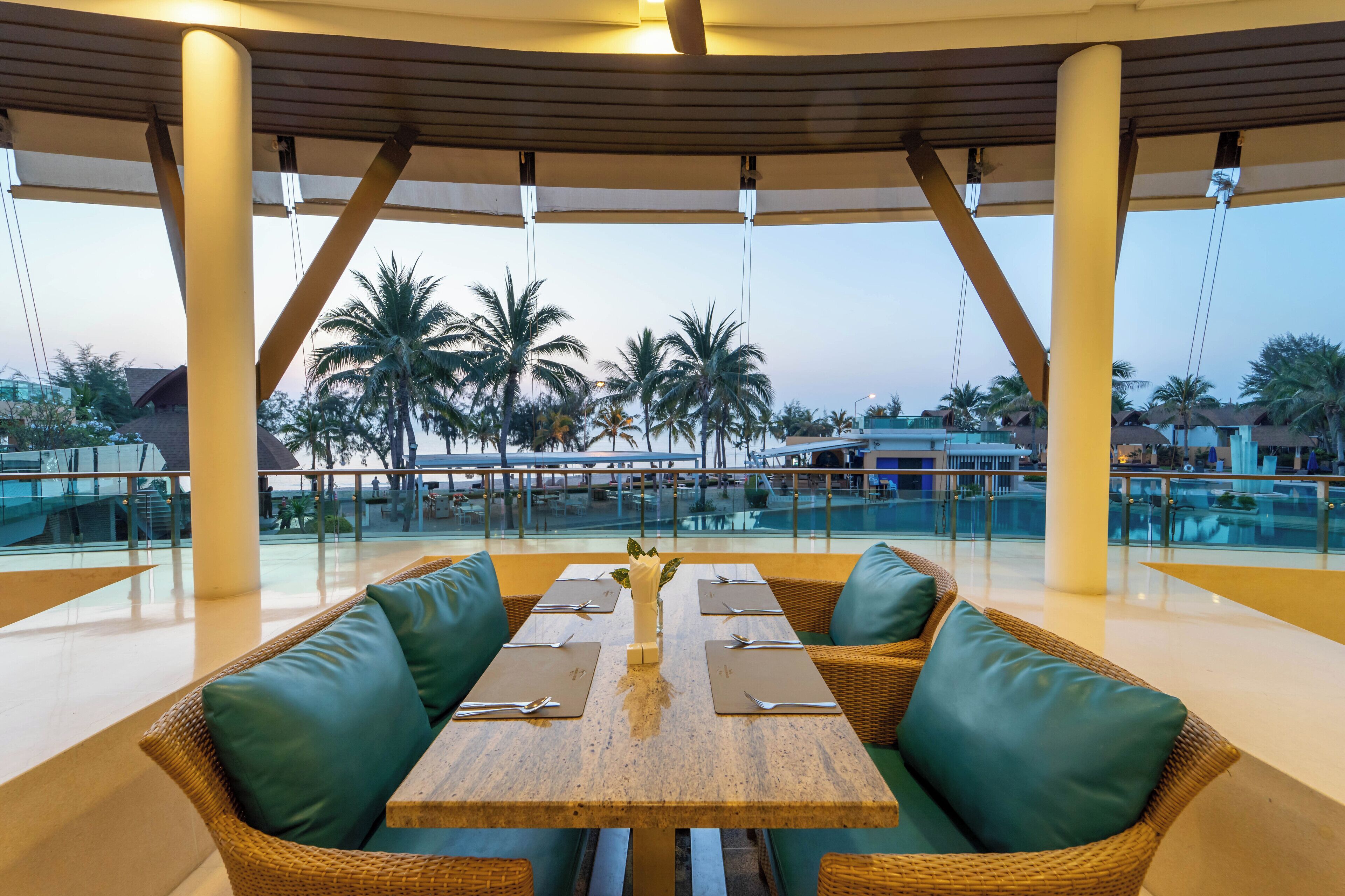 A dining area with a table set for four, surrounded by wicker chairs with green cushions, overlooking a pool and palm trees.