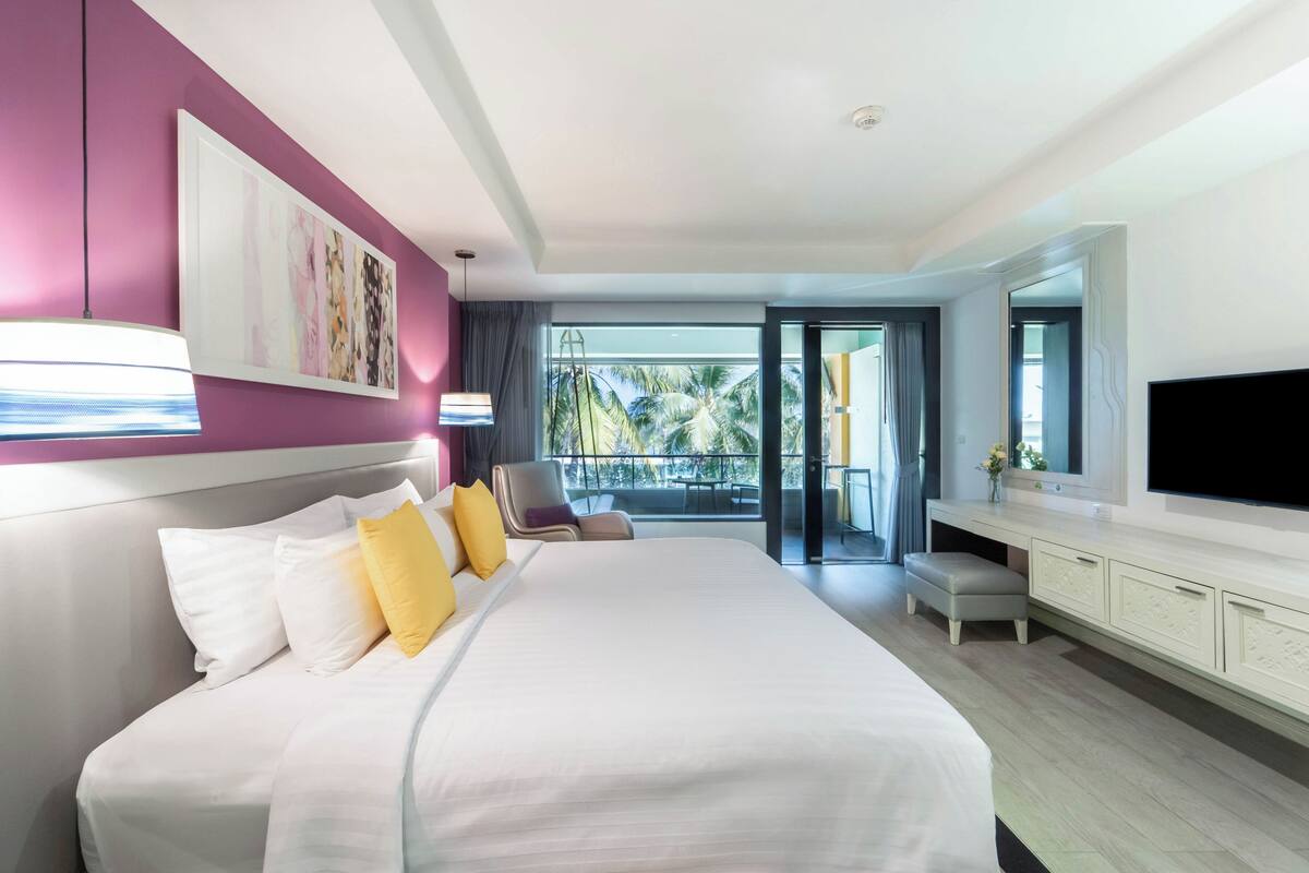 A modern hotel room with a large bed, a flat-screen TV, a balcony with a view of palm trees, and a painting on the wall.
