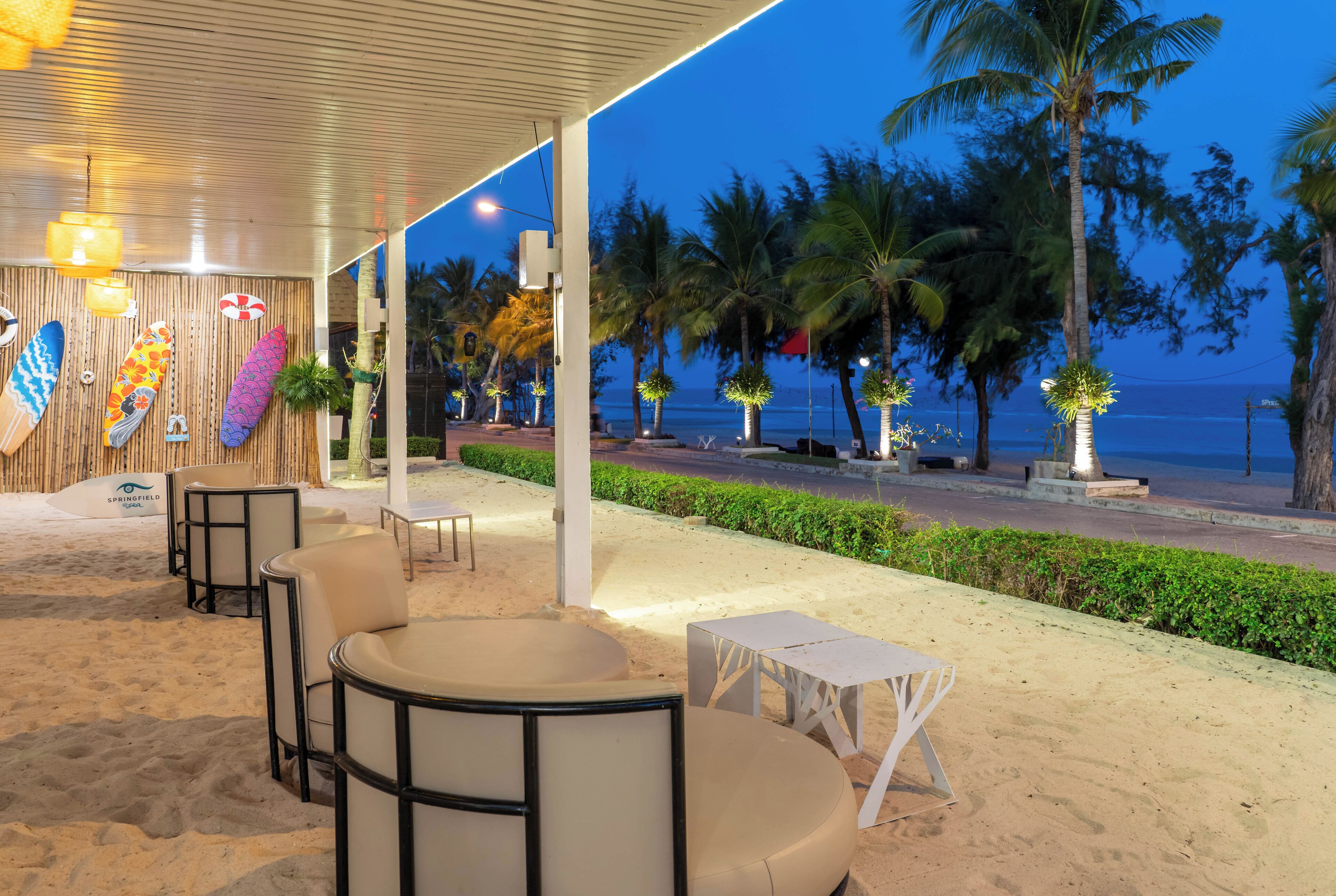 A beachside lounge with a covered seating area, surfboards on display, and a view of the ocean at dusk.