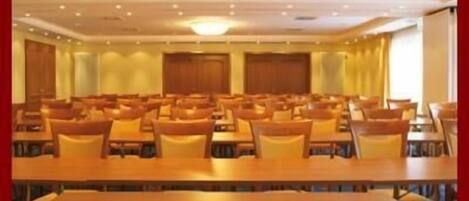 A conference room with rows of tables and chairs, a chandelier, and a large window with curtains.