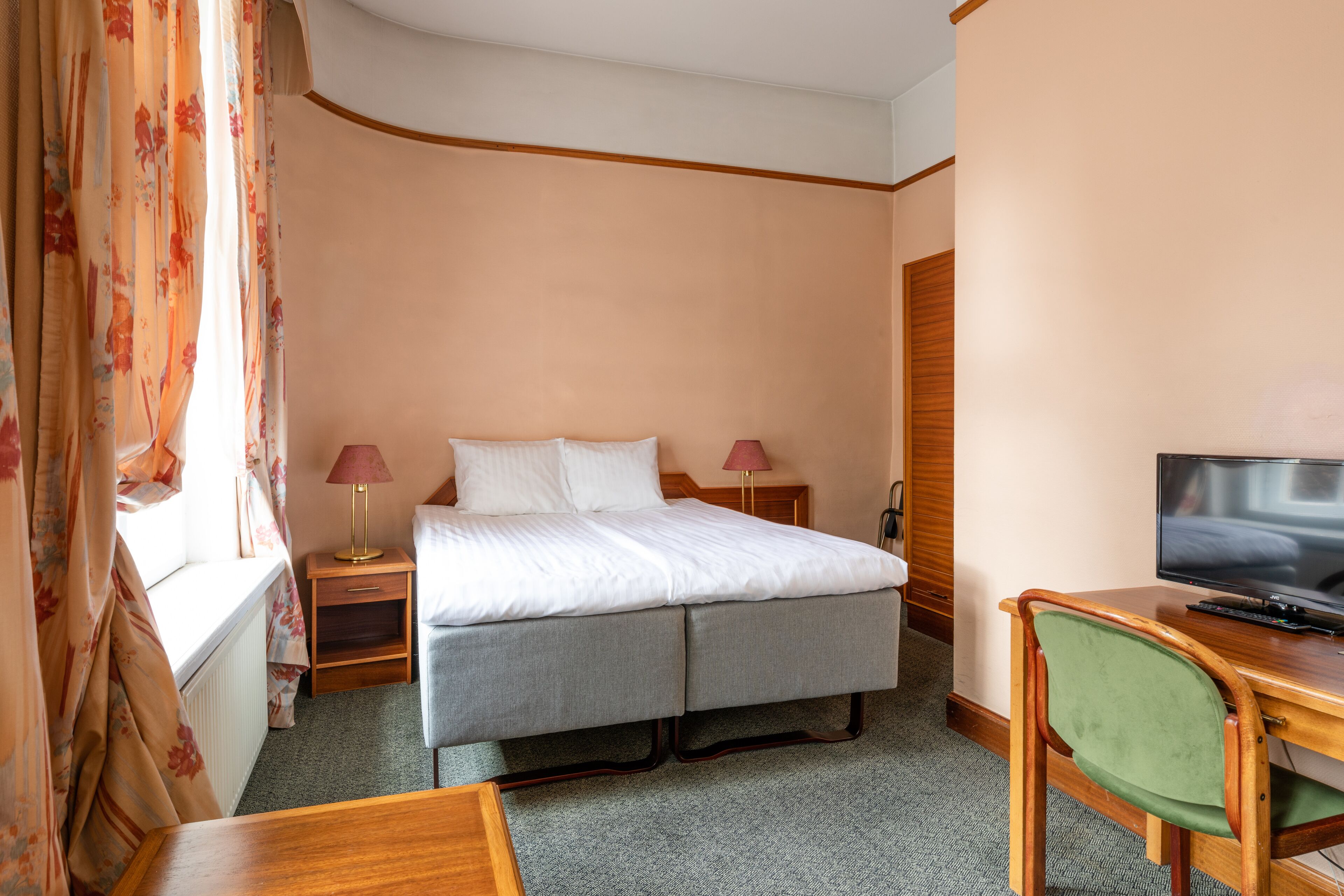 Una camera d'albergo con un letto, una scrivania, una sedia, un televisore e una finestra con tende.
