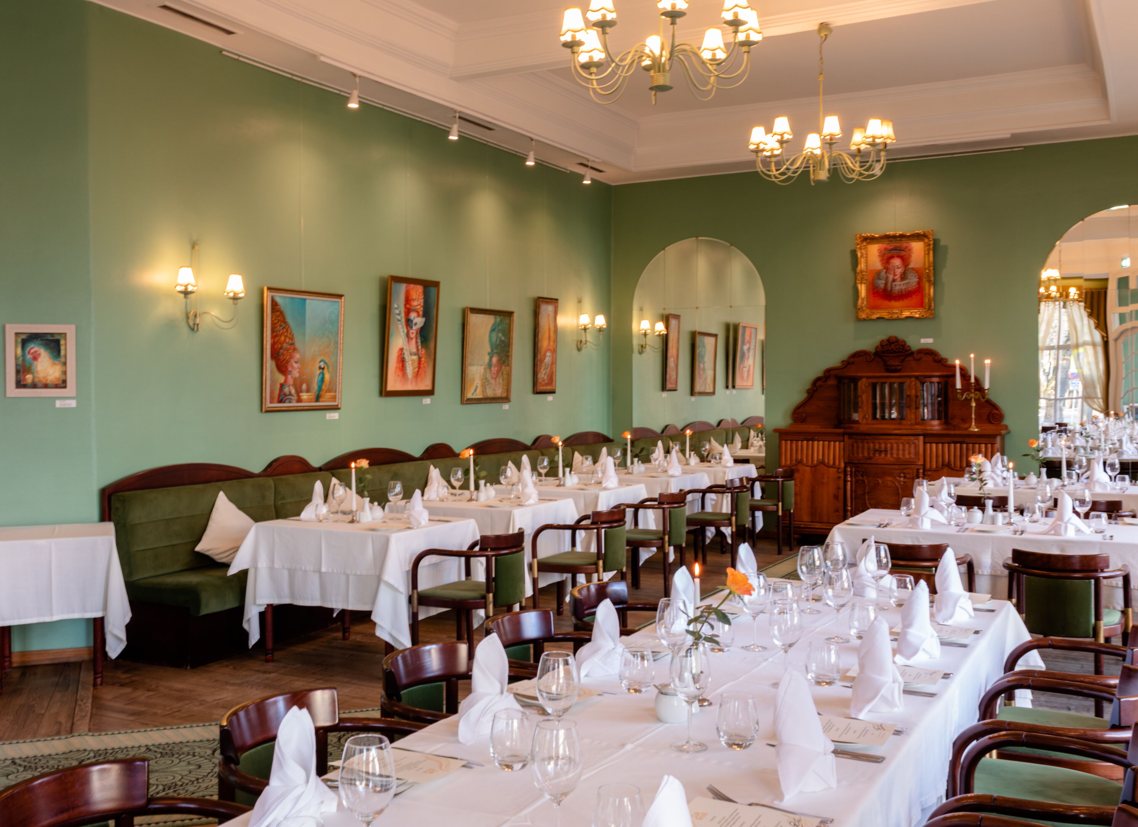 A formal dining room with tables set for service, green upholstered seating, and wall-mounted artwork.