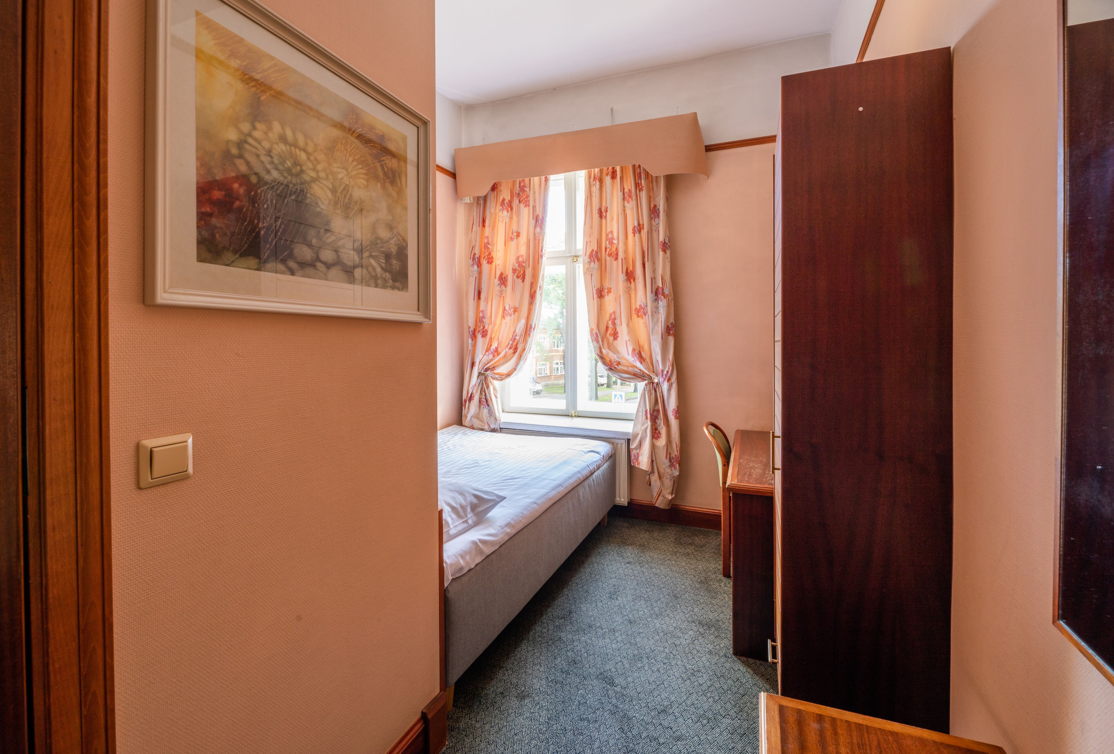 Un dormitorio con una cama, una ventana con cortinas florales, un armario de madera y un cuadro enmarcado en la pared.