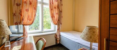 A room with a wooden desk, a bed, a window with floral curtains, and a radiator.