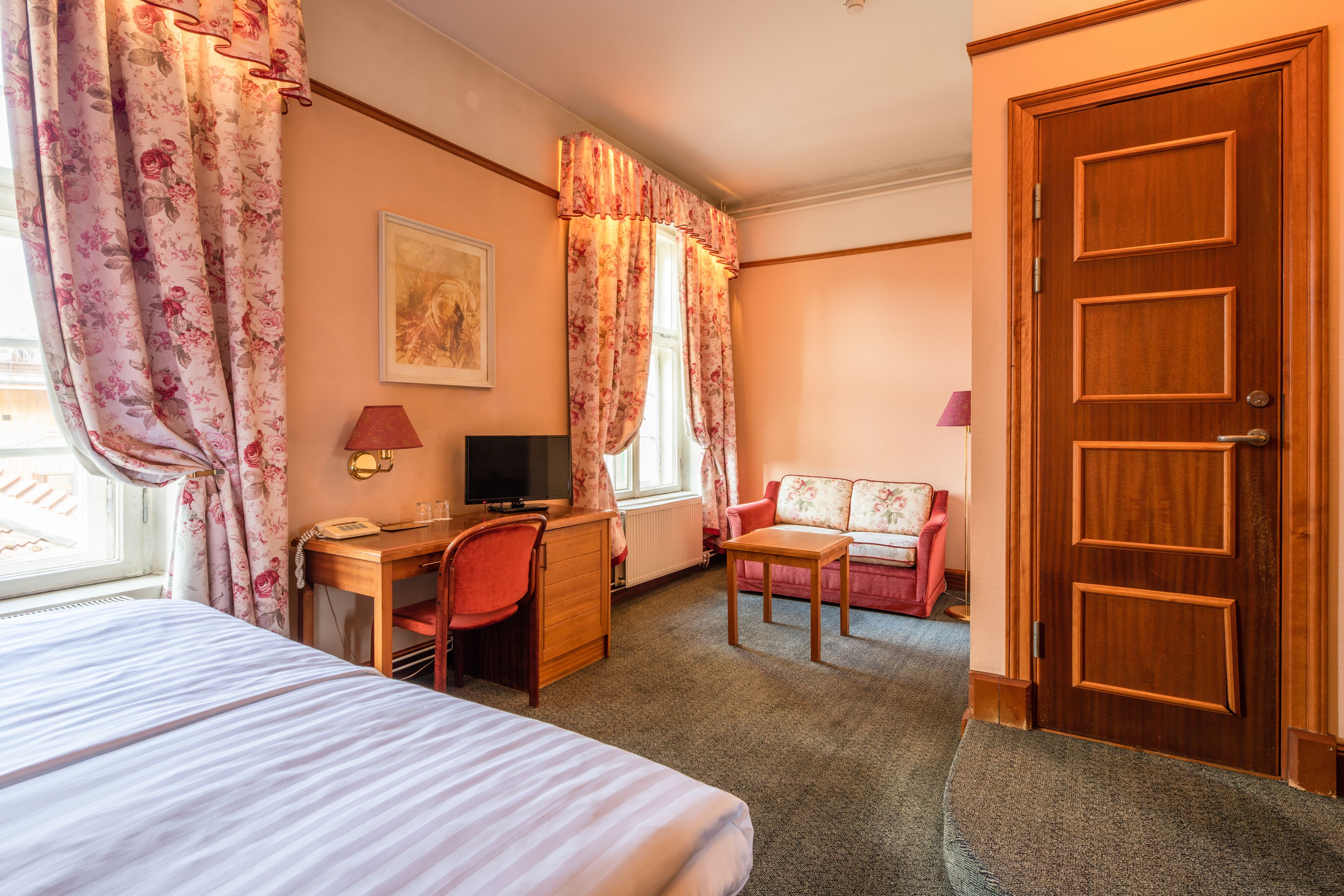 Habitación de hotel con cama, escritorio, computadora, silla, mesita y puerta.