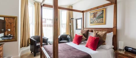 A four-poster bed with a brown bedspread and red and beige pillows, a wooden nightstand, a desk with a lamp, and a painting on the wall.