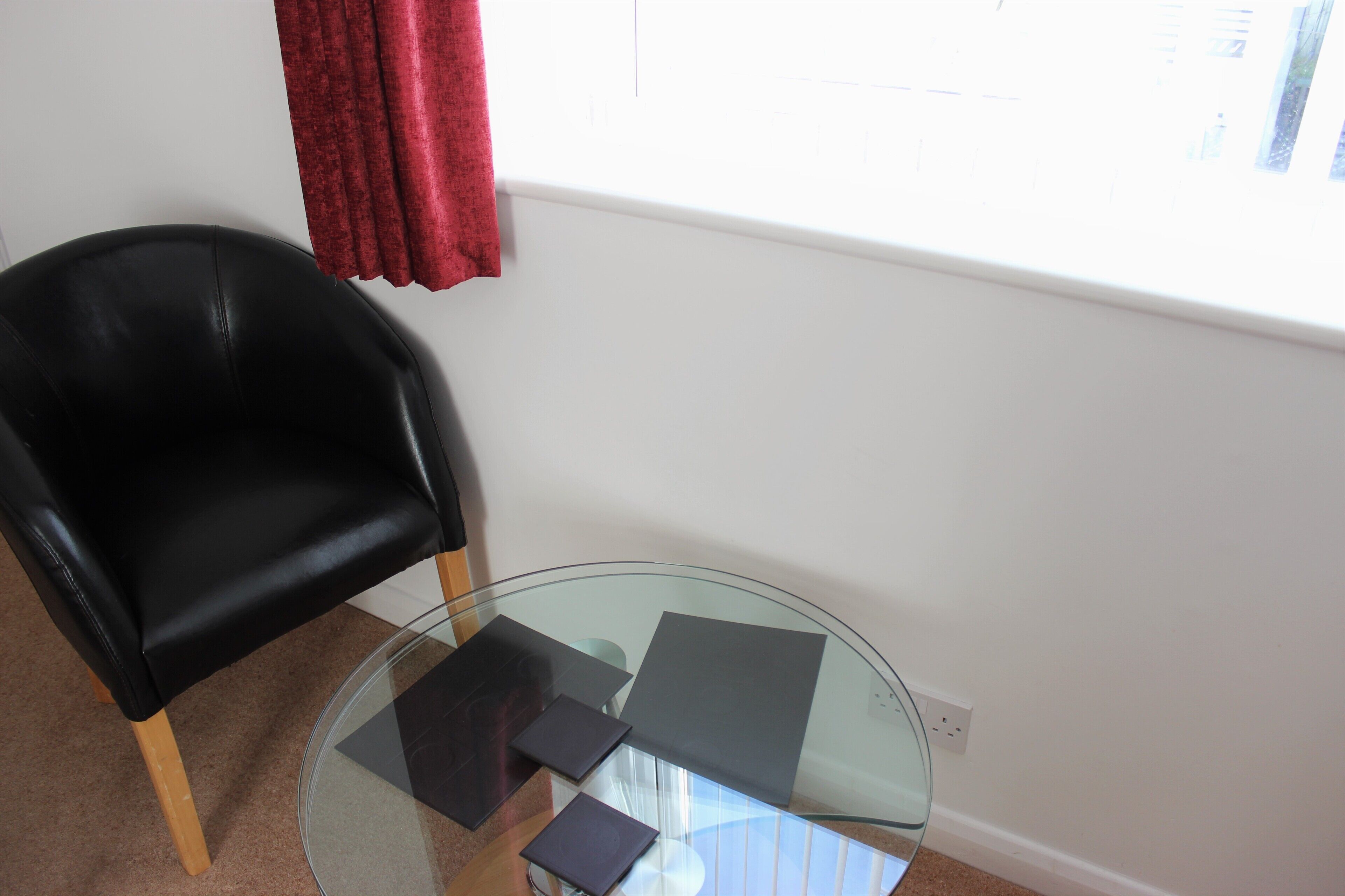 Una habitación con una mesa de vidrio, una silla negra y una ventana con cortina roja.