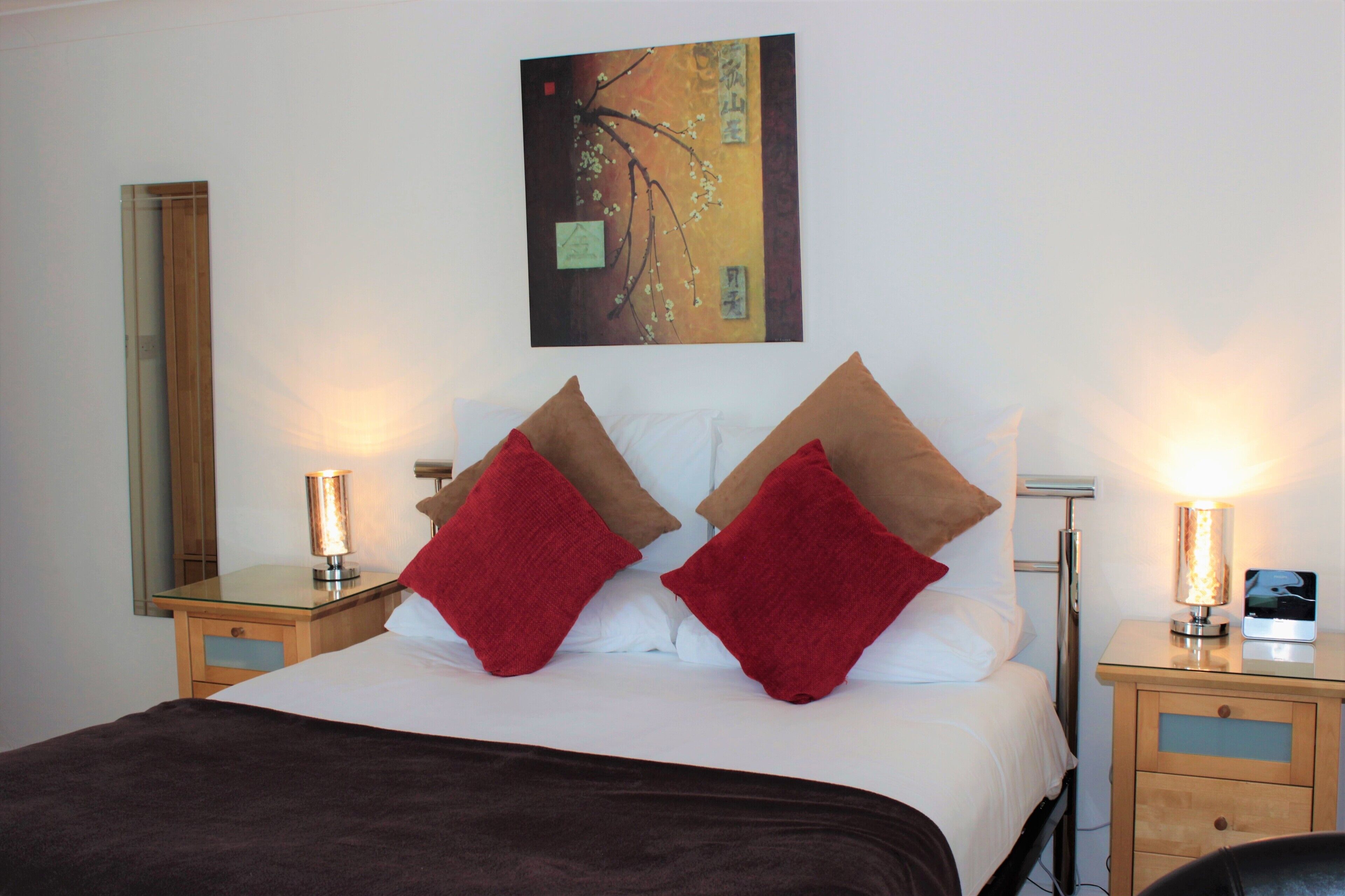 A neatly made bed with red and brown pillows, a bedside table with a lamp, and a wall-mounted painting.