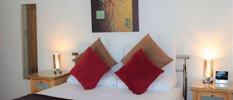 A neatly made bed with red and brown pillows, a bedside table with a lamp, and a wall-mounted painting.