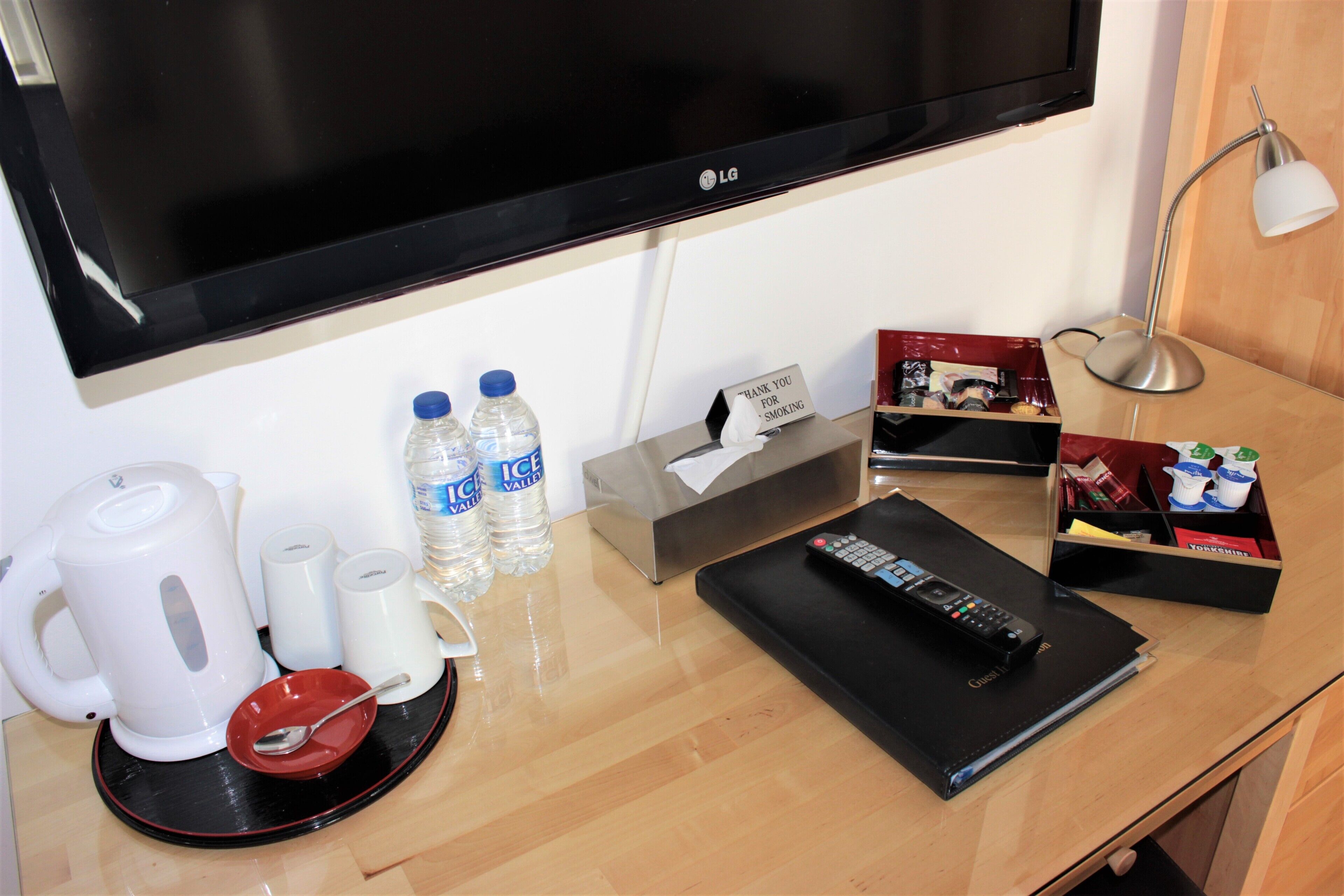 A hotel room with a flat-screen TV, a kettle, water bottles, a remote control, and a tray with a spoon.