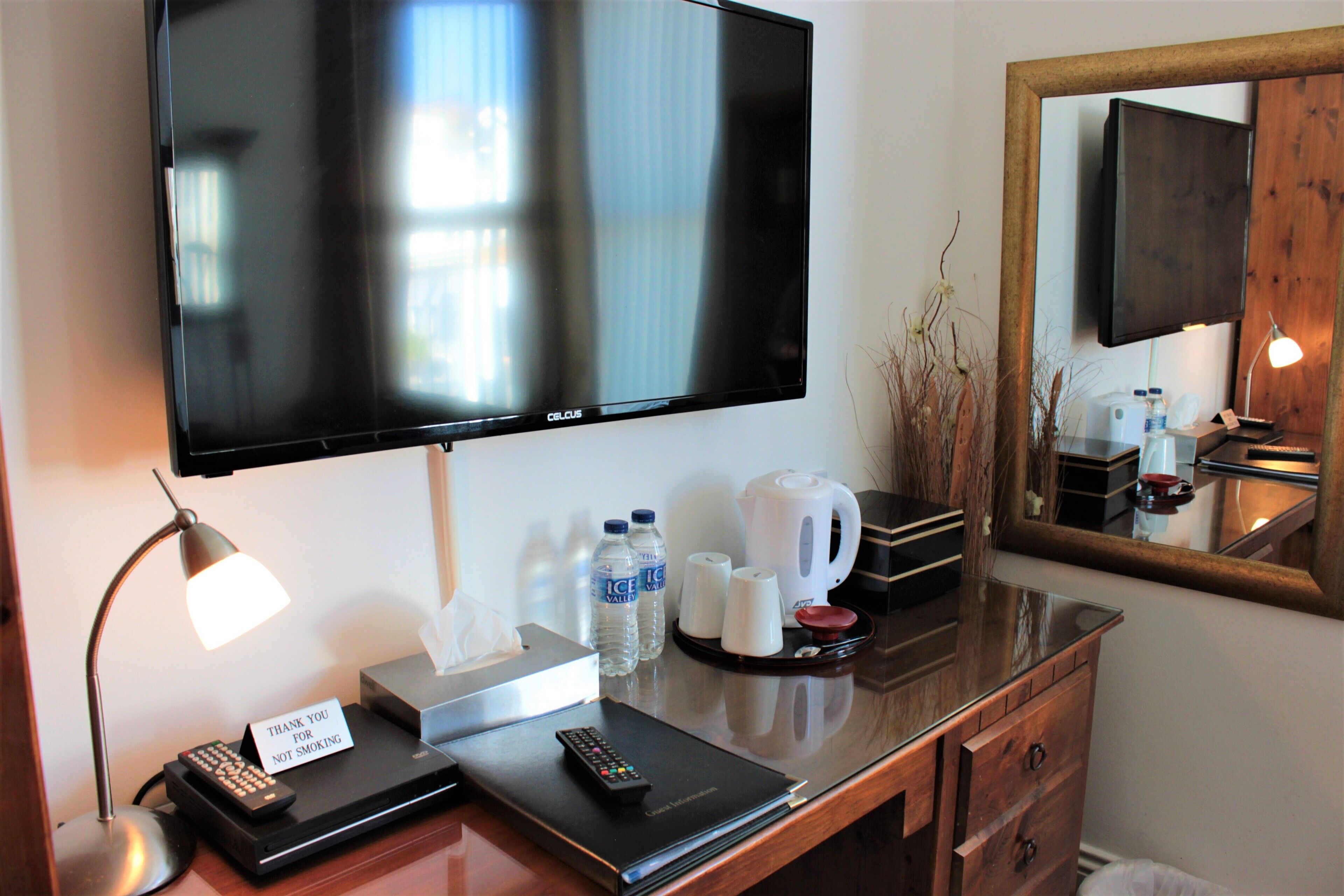 A hotel room with a flat-screen TV, a desk with a lamp, a remote control, a notepad, and a sign.