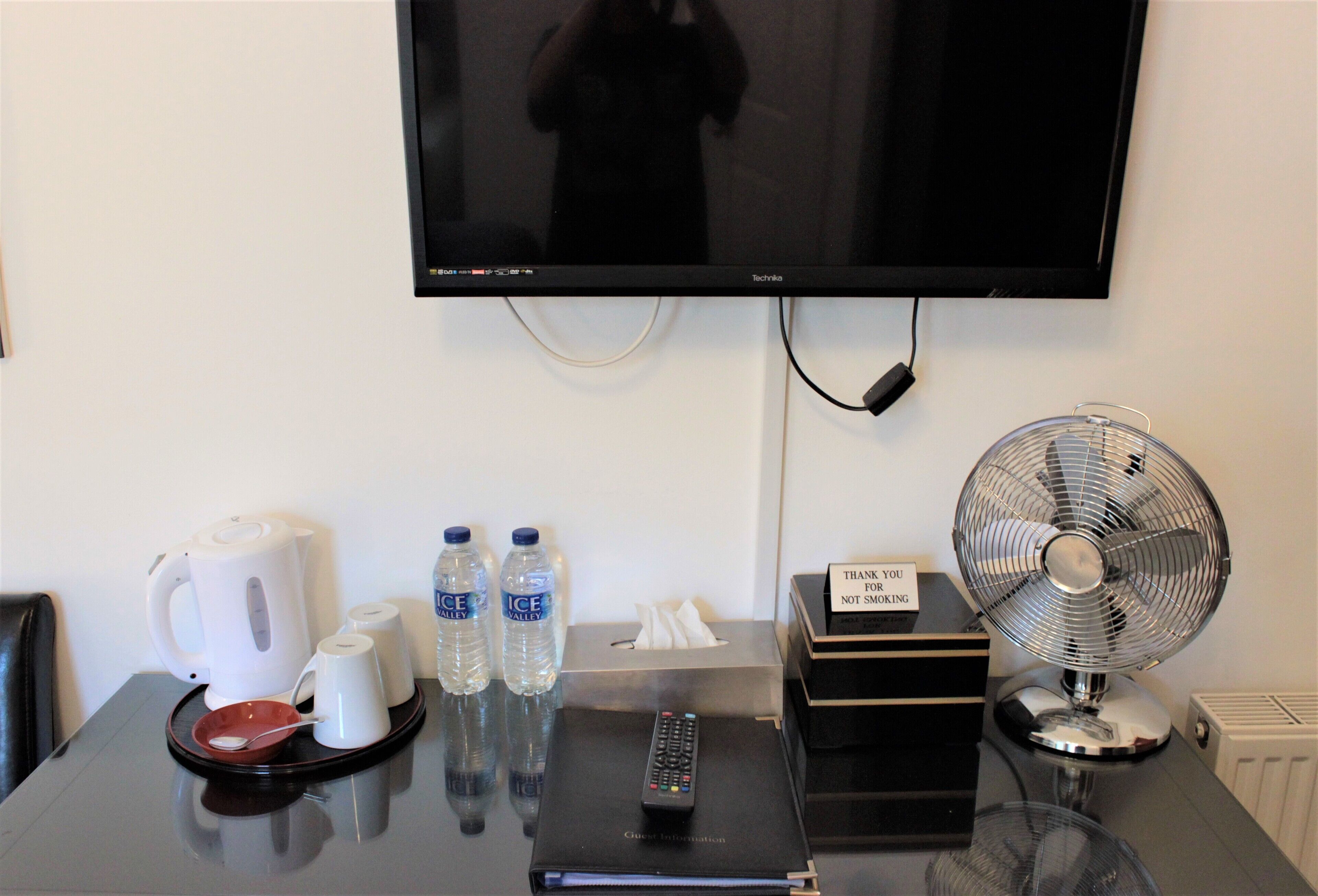 A hotel room with a flat-screen TV mounted on the wall, a table with a kettle, two water bottles, a remote control, a fan, and a sign that says "Thank you for not smoking."