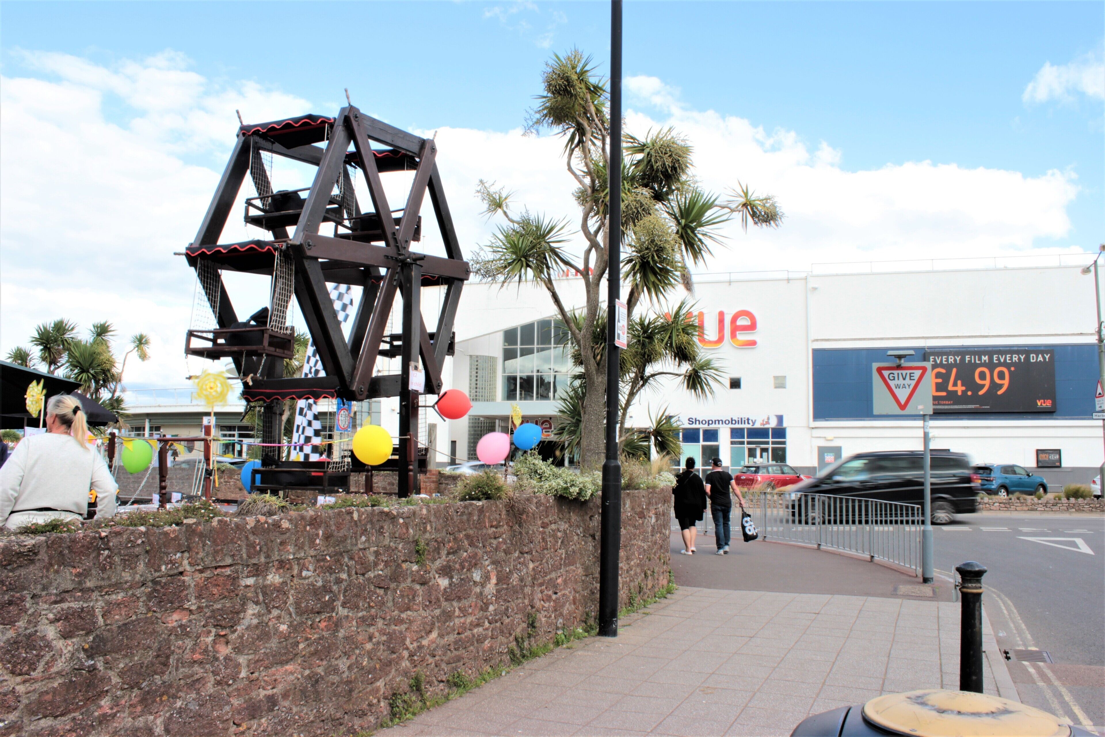 A public square with a large metal sculpture, palm trees, and a building with a large sign advertising a film rental service.