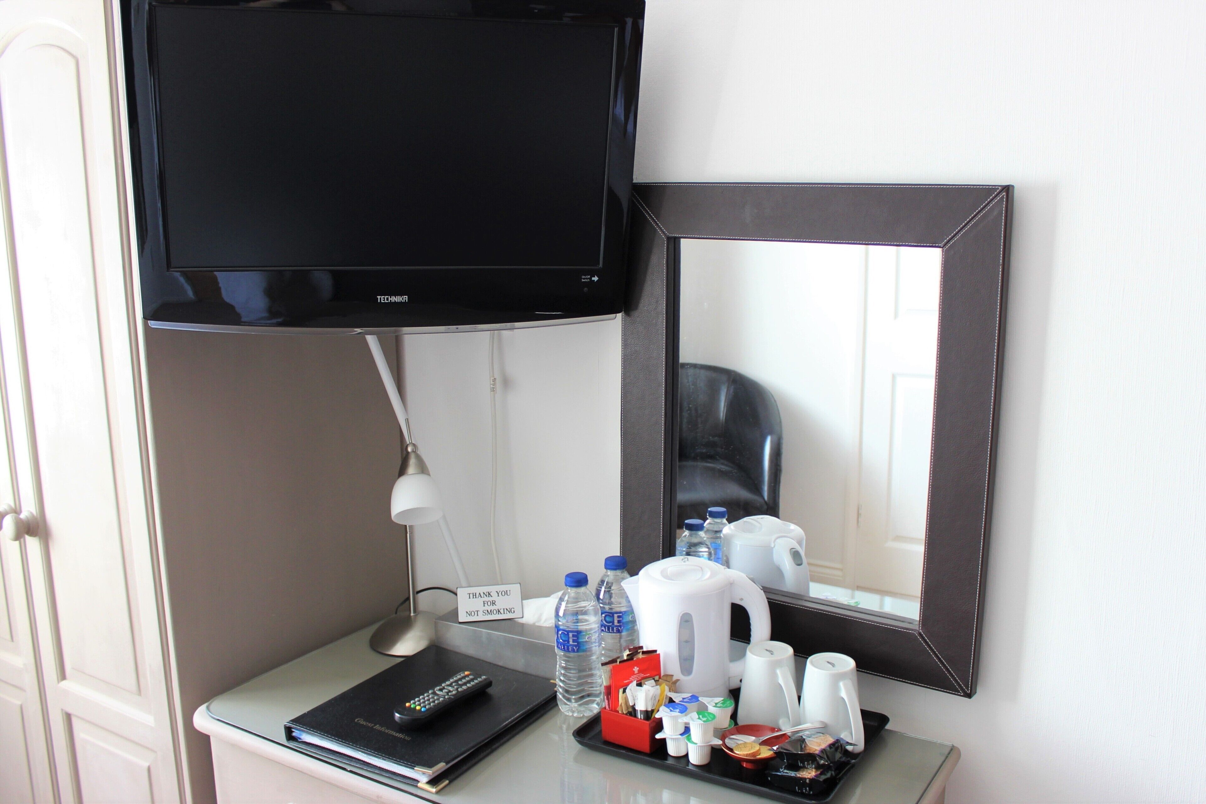 Hotelkamer met een wandgemonteerde flatscreen-tv, een nachtkastje met een afstandsbediening, een waterfles, een koffiezetapparaat en een dienblad met diverse voorwerpen.