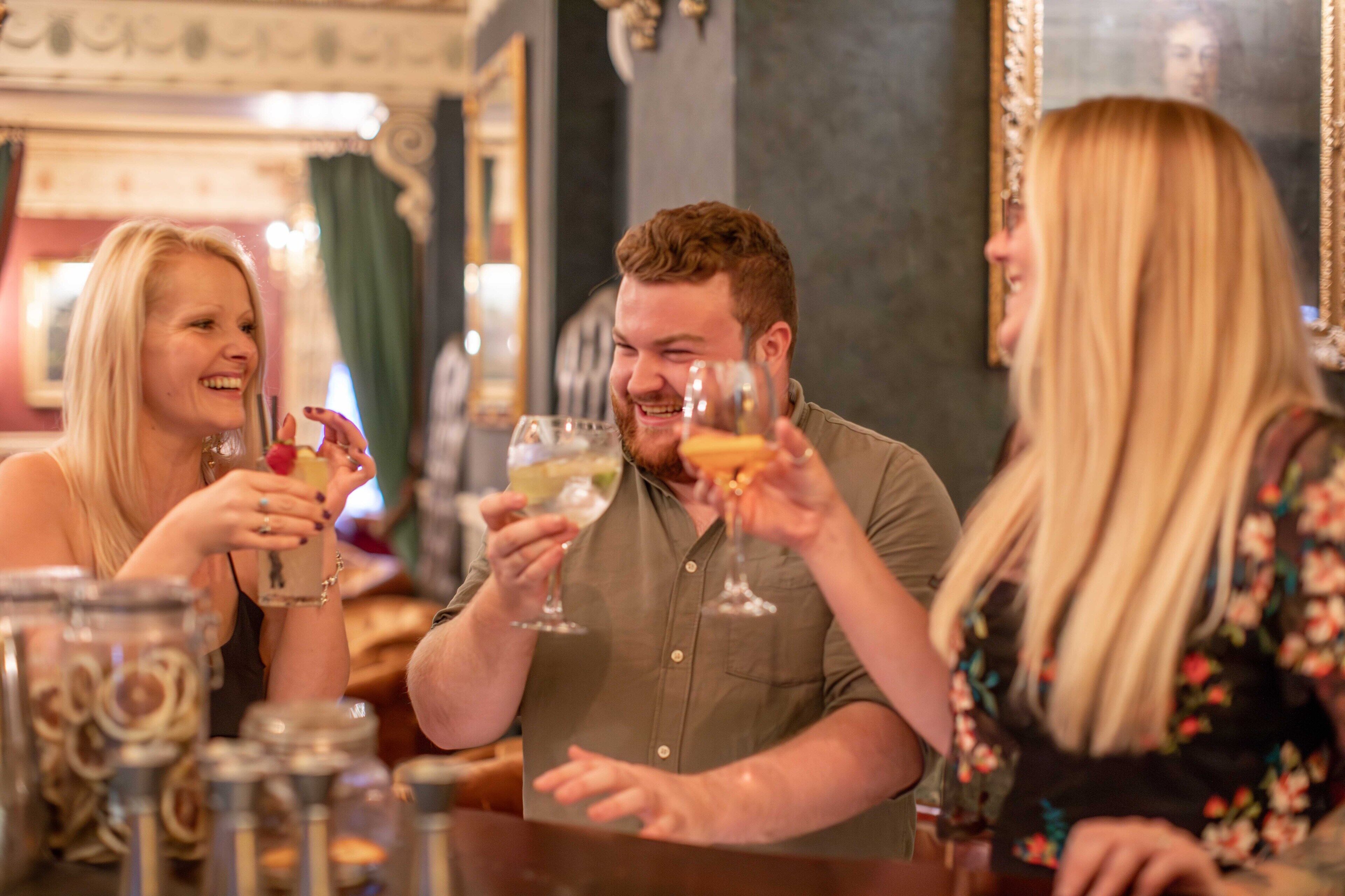 A group of people toasting with drinks at a bar with ornate decor and a mirror reflecting part of the scene.