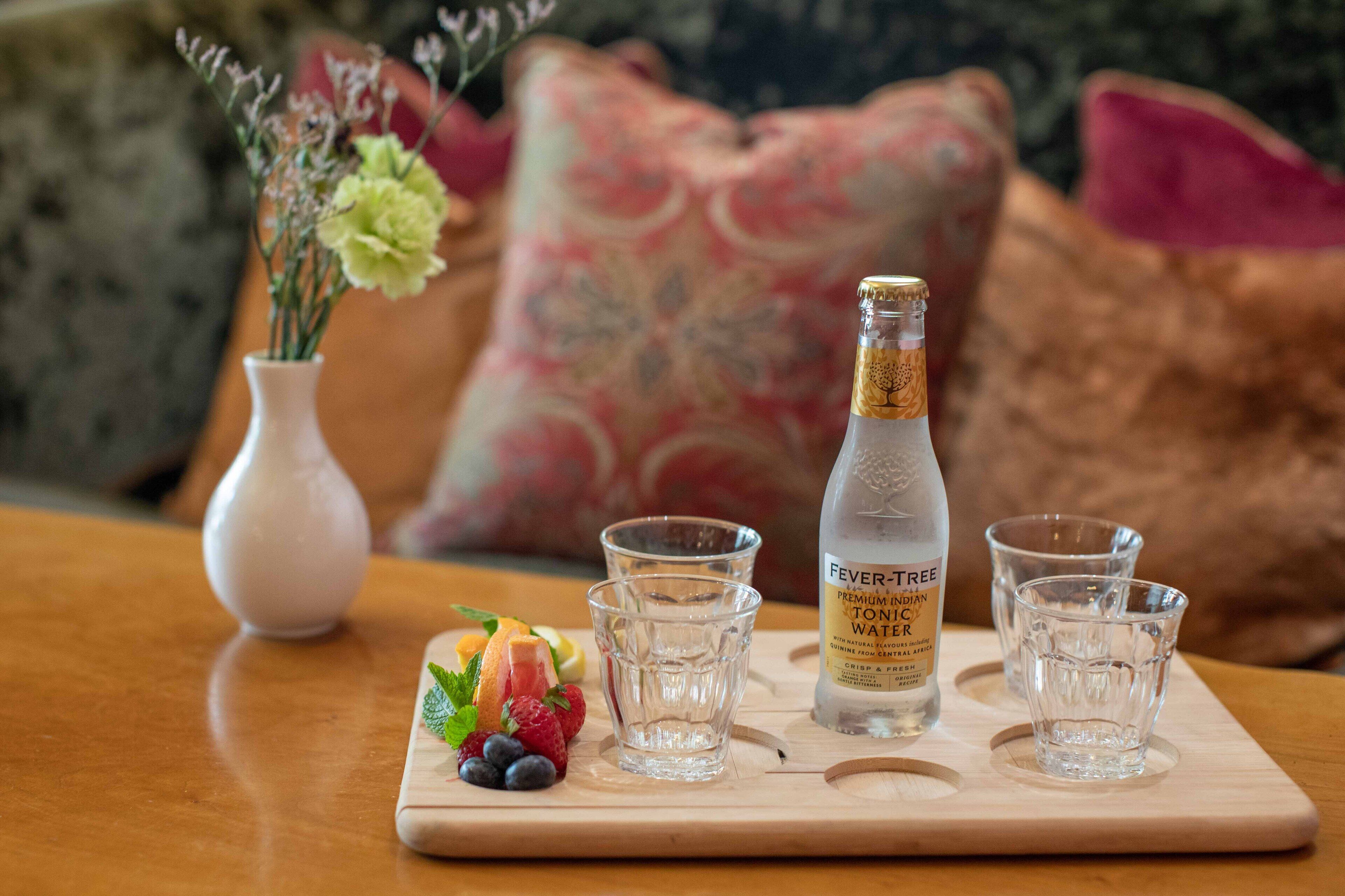 A wooden tray with a bottle of Fever Tree Premium Indian Tonics Water, three shot glasses, and a variety of fresh fruits.