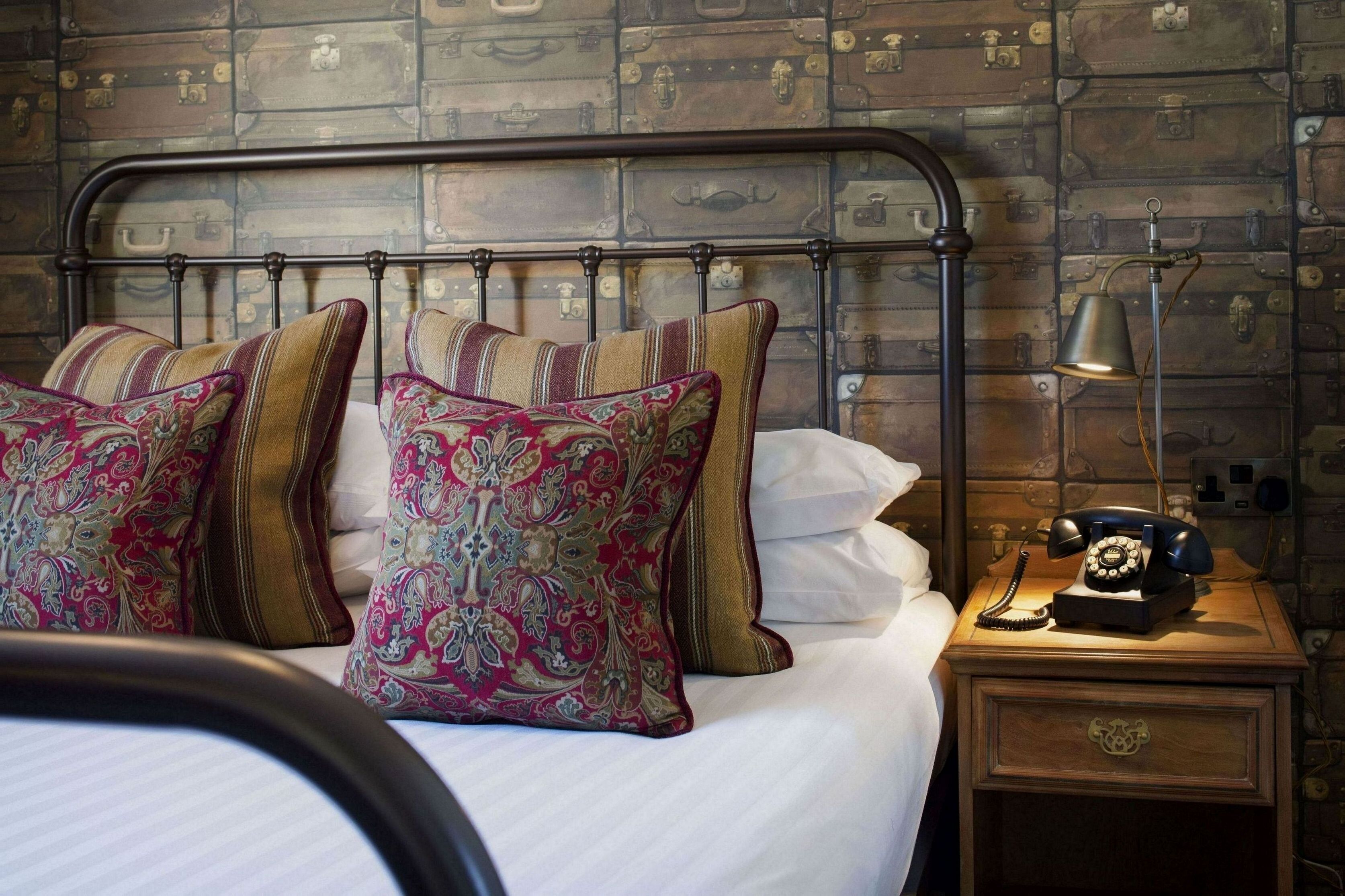 A bed with patterned pillows, a metal headboard, a vintage telephone, and a wall with a suitcase motif.