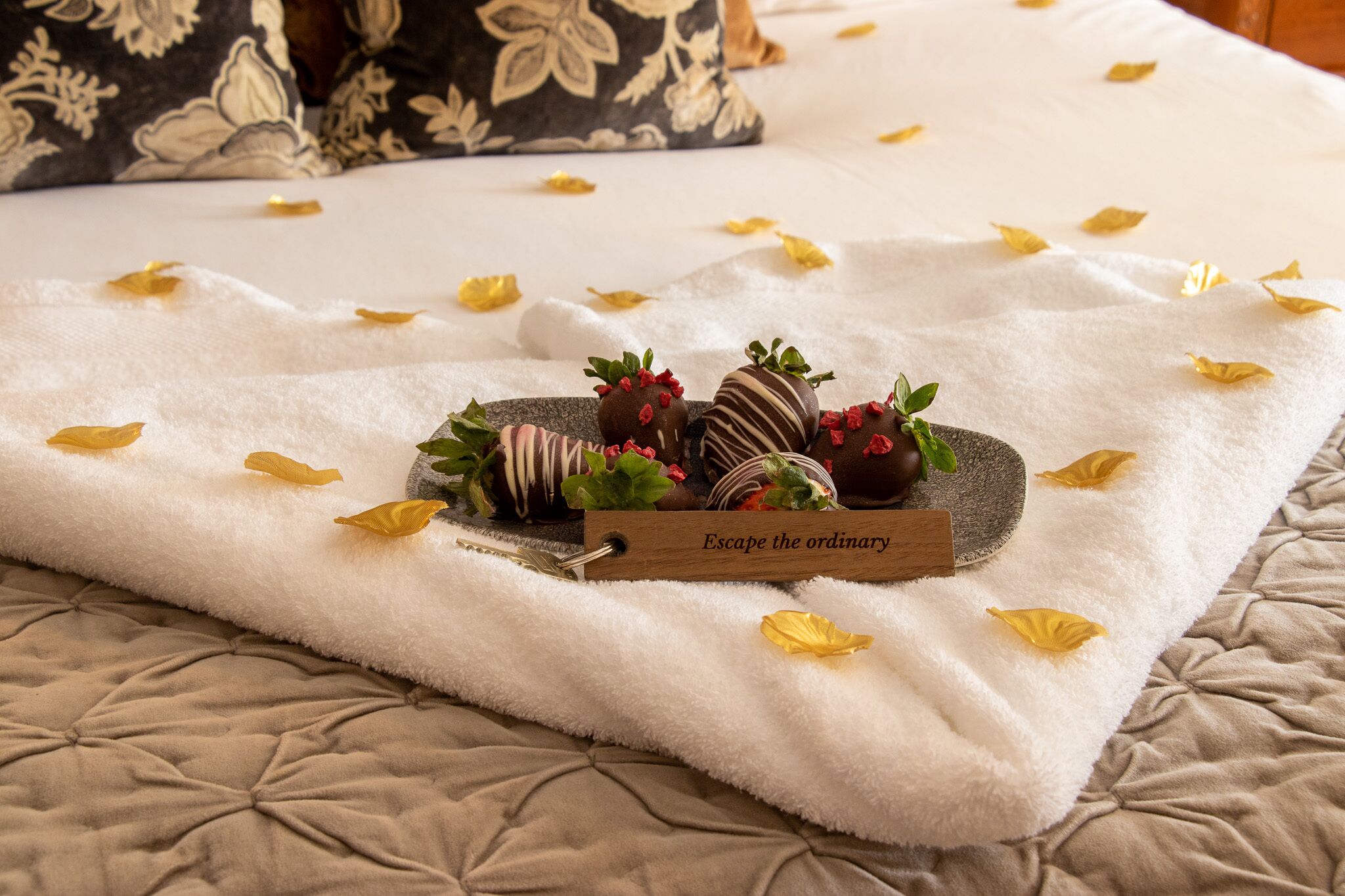 A neatly arranged tray of chocolate-covered strawberries and a wooden sign on a bed with a white towel and scattered yellow petals.