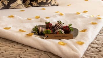 A neatly arranged tray of chocolate-covered strawberries and a wooden sign on a bed with a white towel and scattered yellow petals.