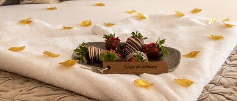 Bandeja organizada com morangos cobertos de chocolate e uma placa de madeira sobre a cama, com uma toalha branca e pétalas amarelas espalhadas.
