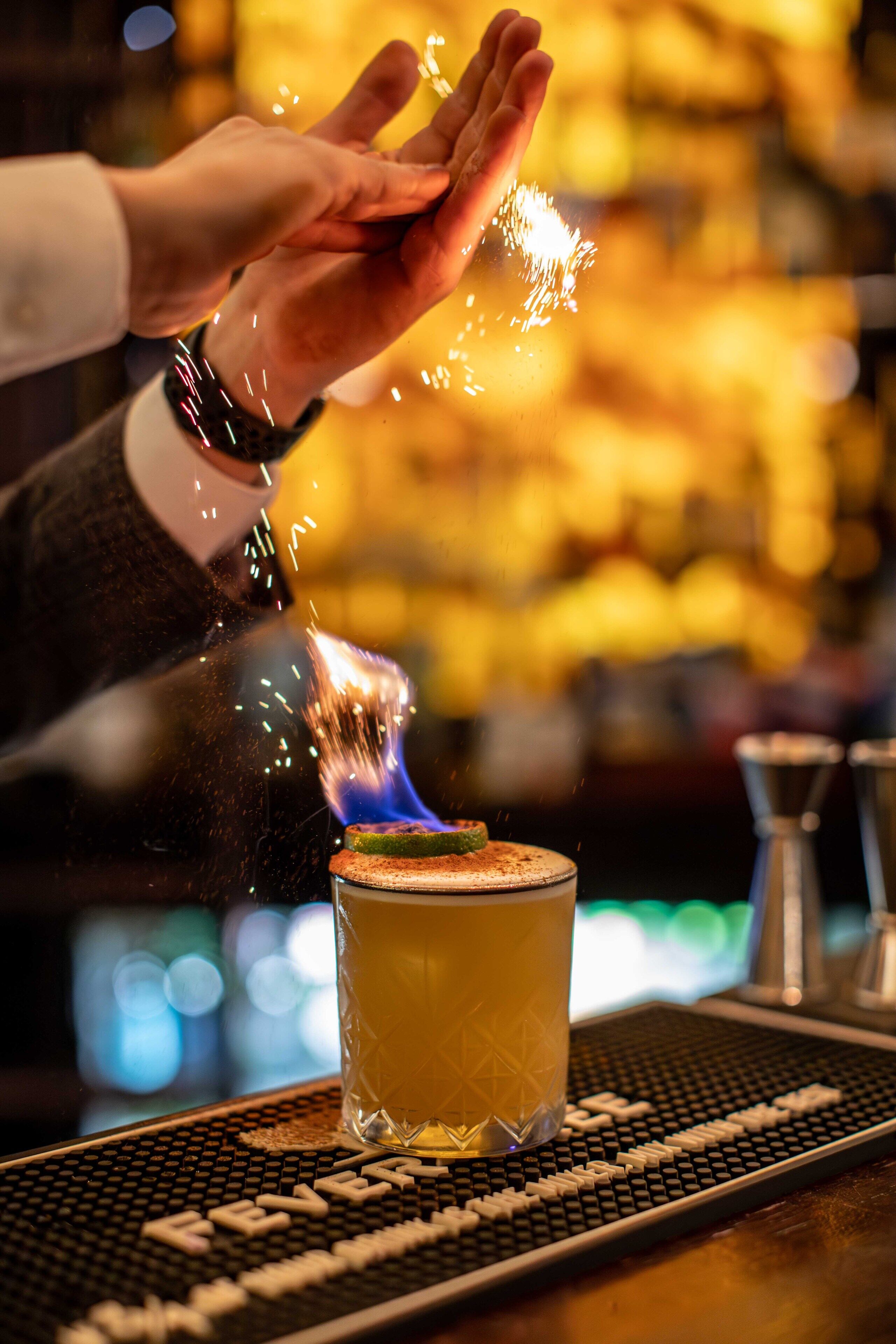 A bartender pouring a drink with a lit match into a glass on a bar mat.