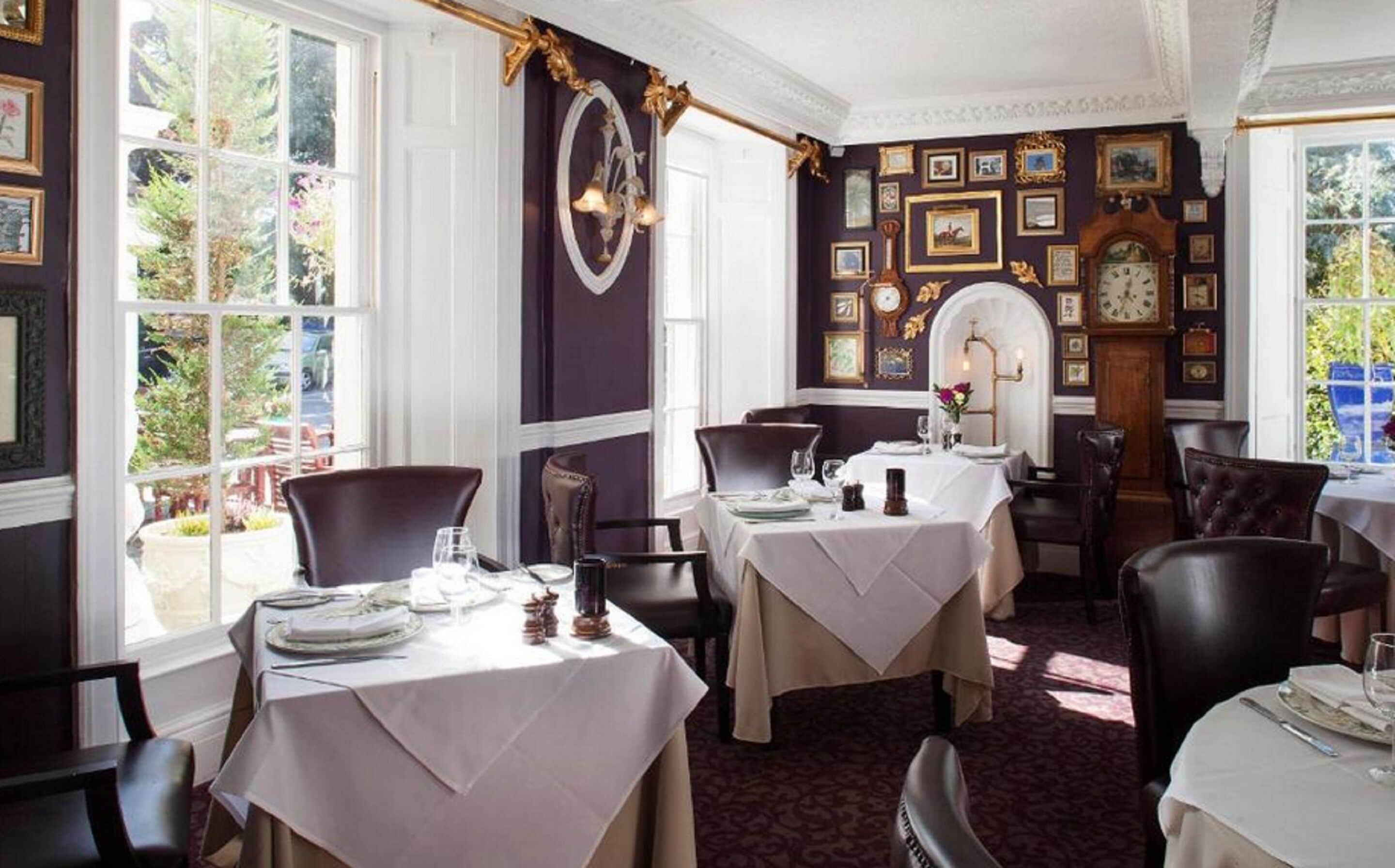 Comedor con mesas dispuestas para dos, ventanas amplias y una pared decorada con cuadros y un reloj.