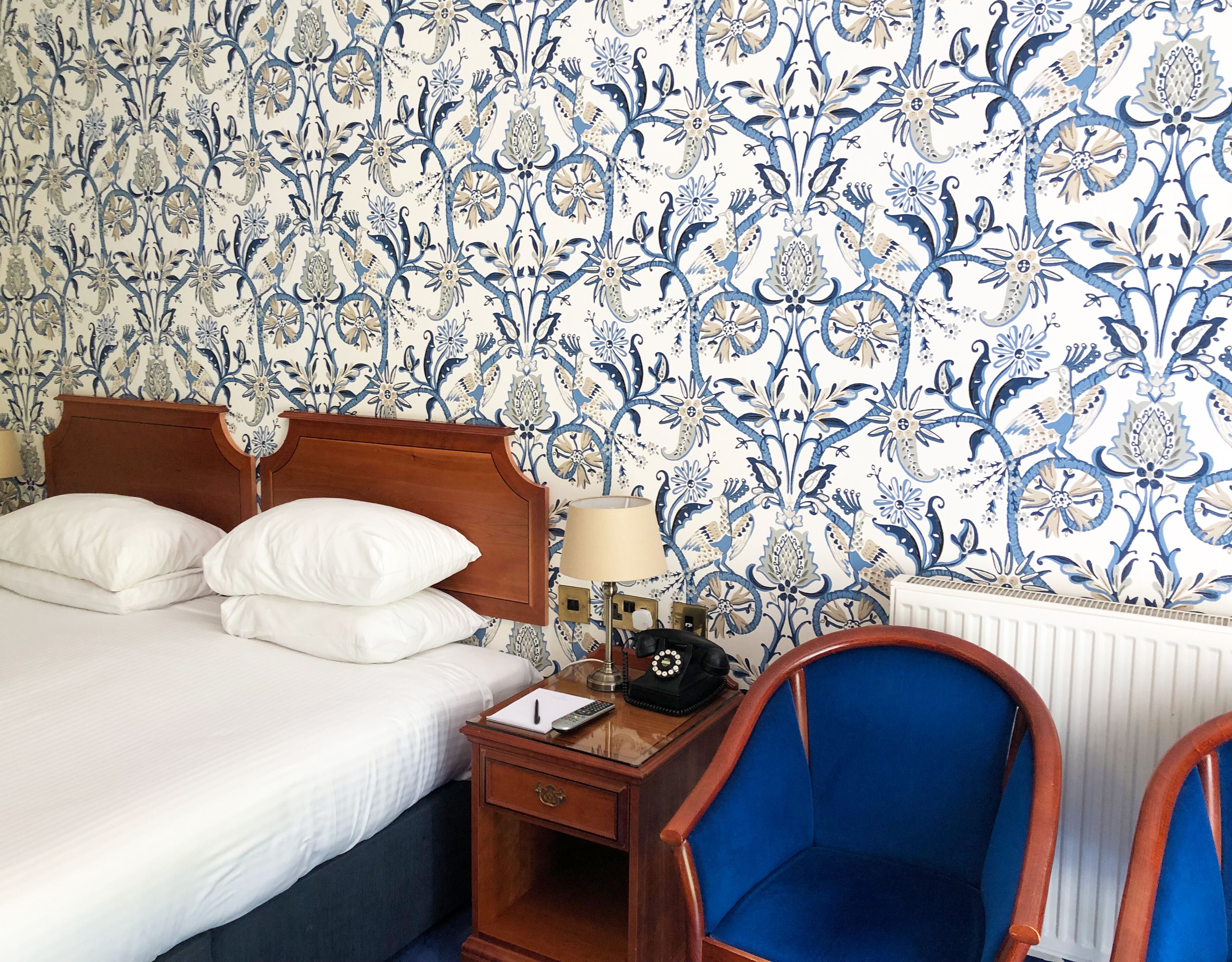 A hotel room with a bed, bedside table, lamp, chair, and a patterned wallpaper.