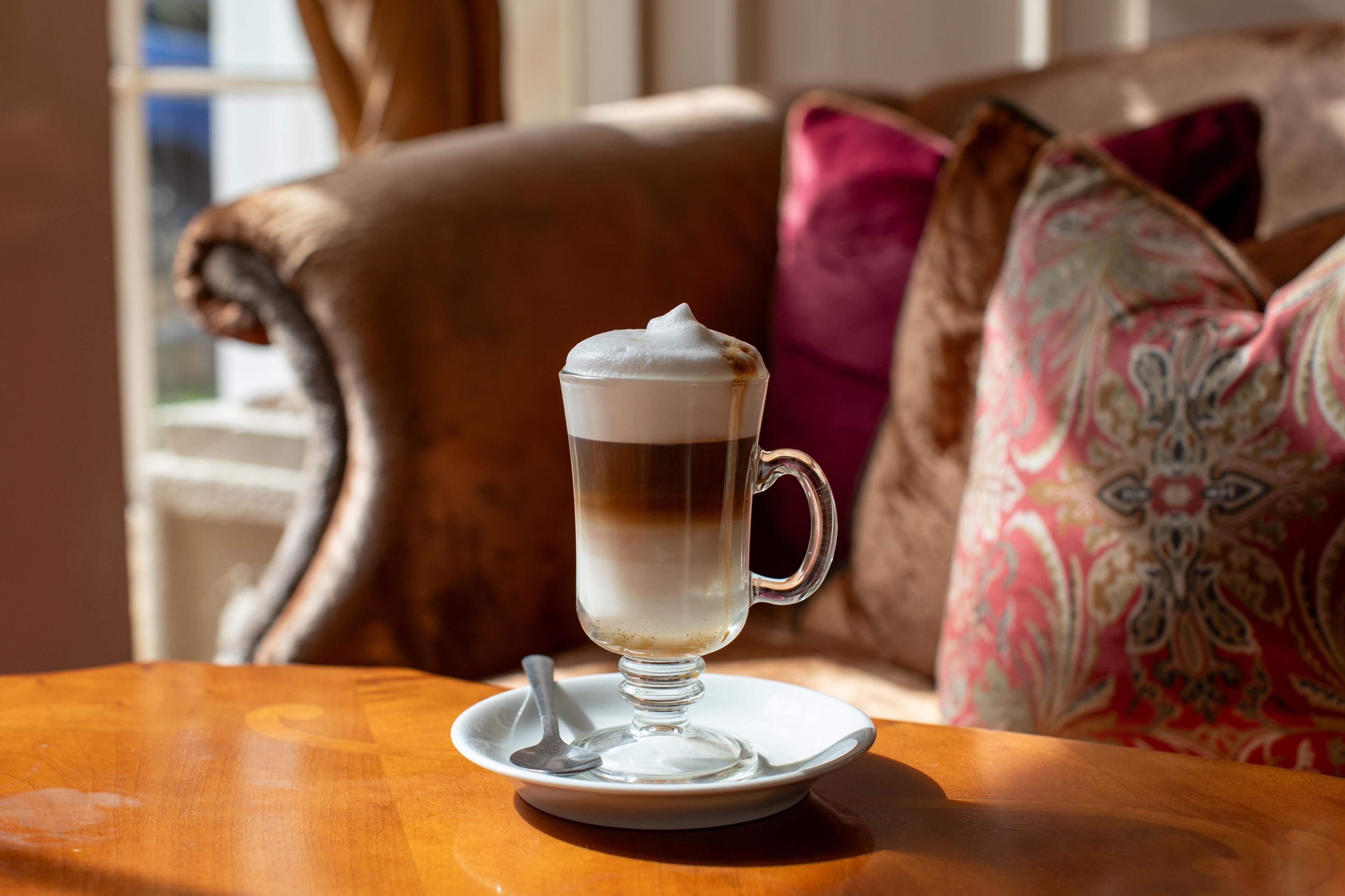 A cup of coffee on a saucer with a spoon, placed on a wooden table in a room with a brown leather couch and patterned pillows.