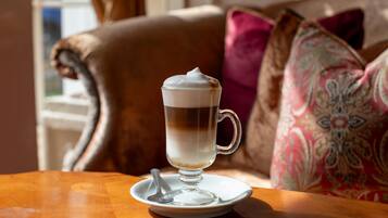 A cup of coffee on a saucer with a spoon, placed on a wooden table in a room with a brown leather couch and patterned pillows.