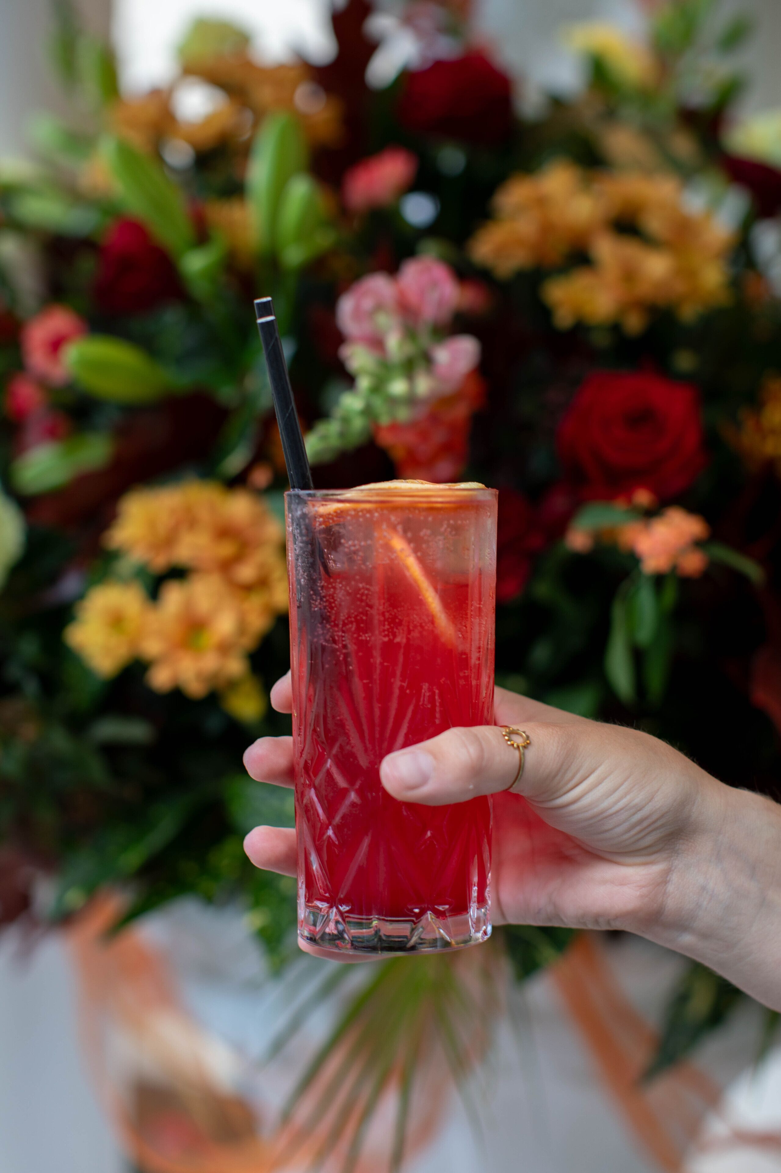 A hand holding a tall glass with a red beverage and a straw, set against a floral background.
