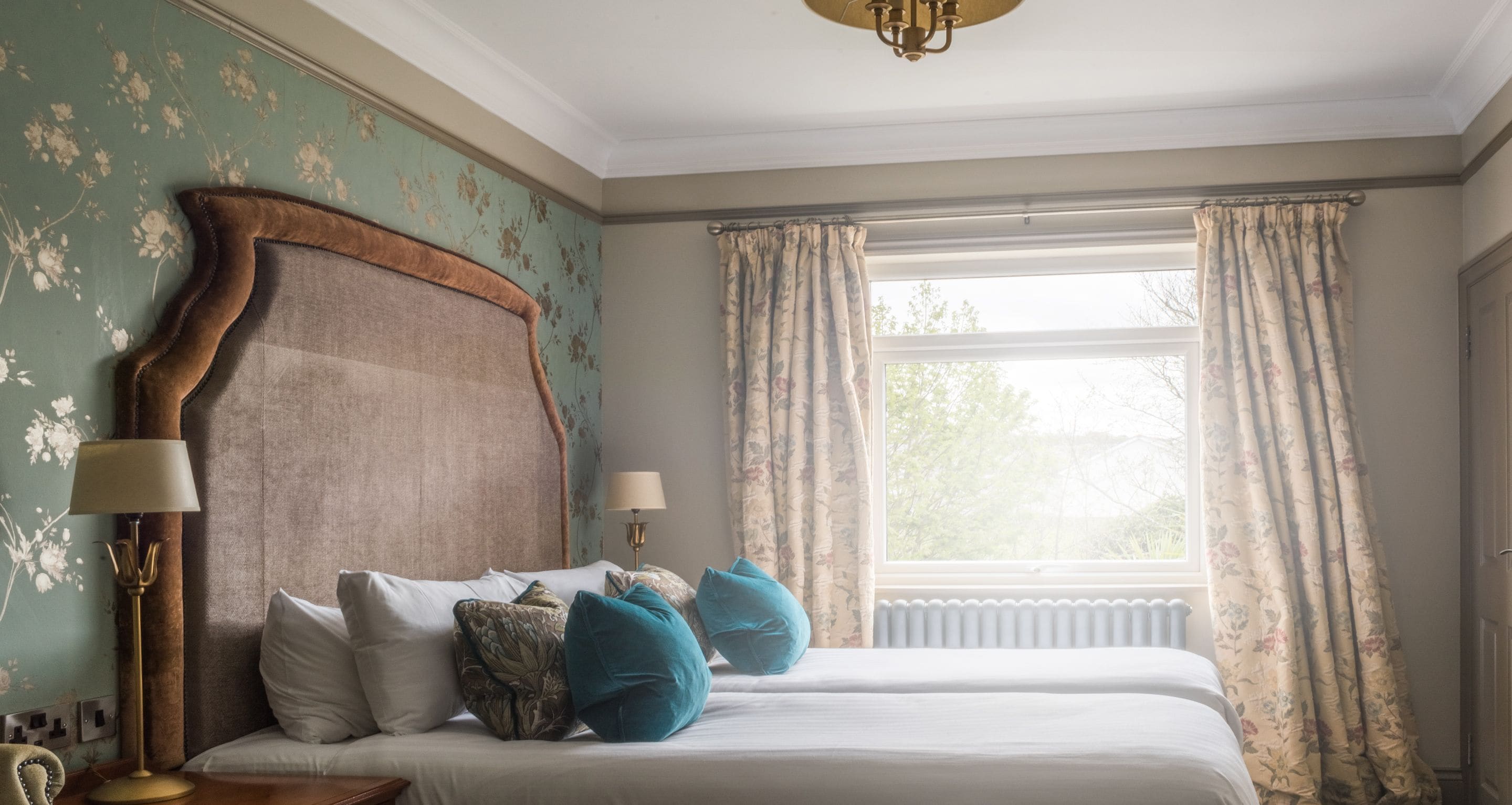 A bedroom with a large bed, two bedside lamps, floral wallpaper, and a window with curtains.