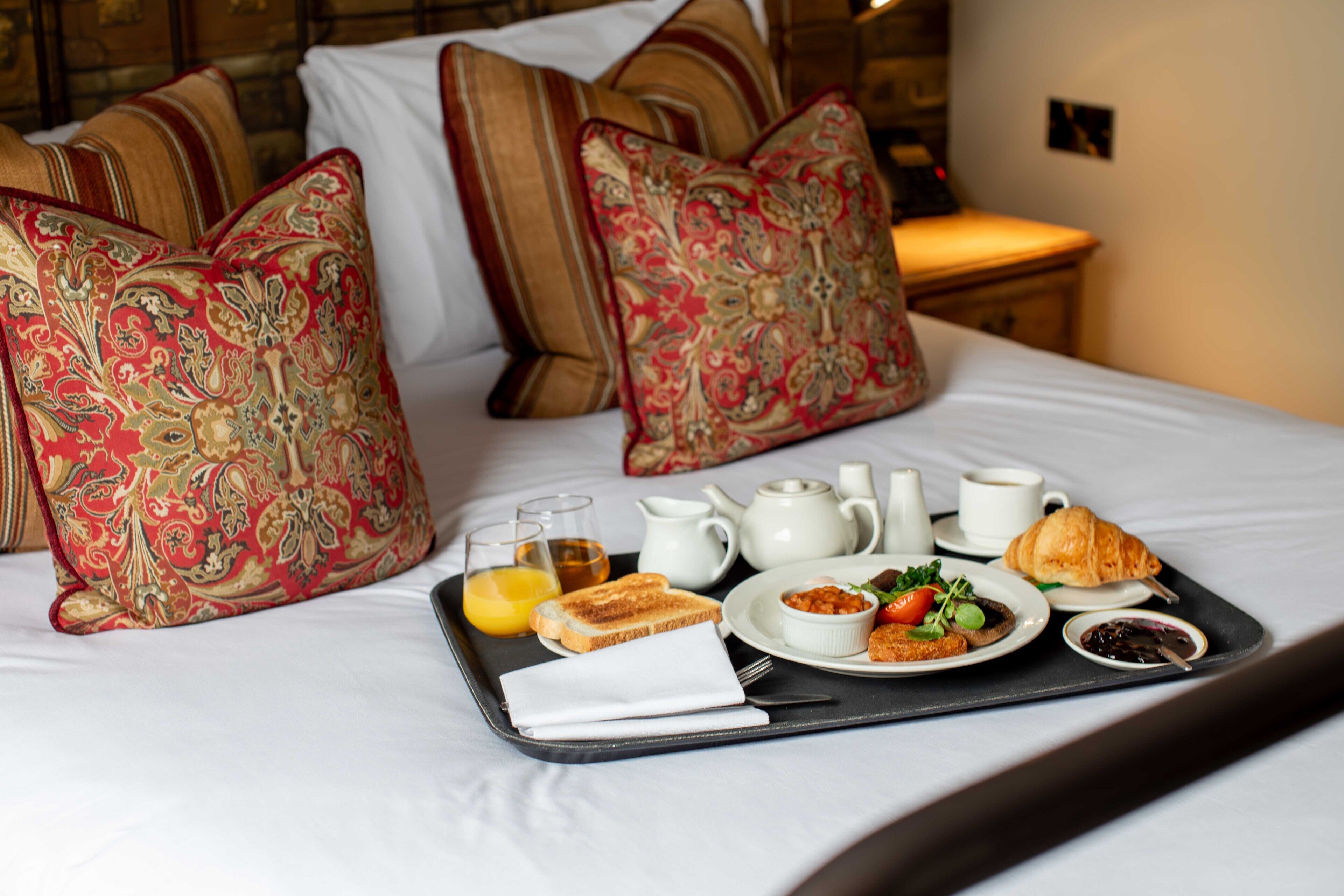 A neatly made bed with decorative pillows, a tray with breakfast items including toast, eggs, and a croissant, and a bedside table with a lamp.