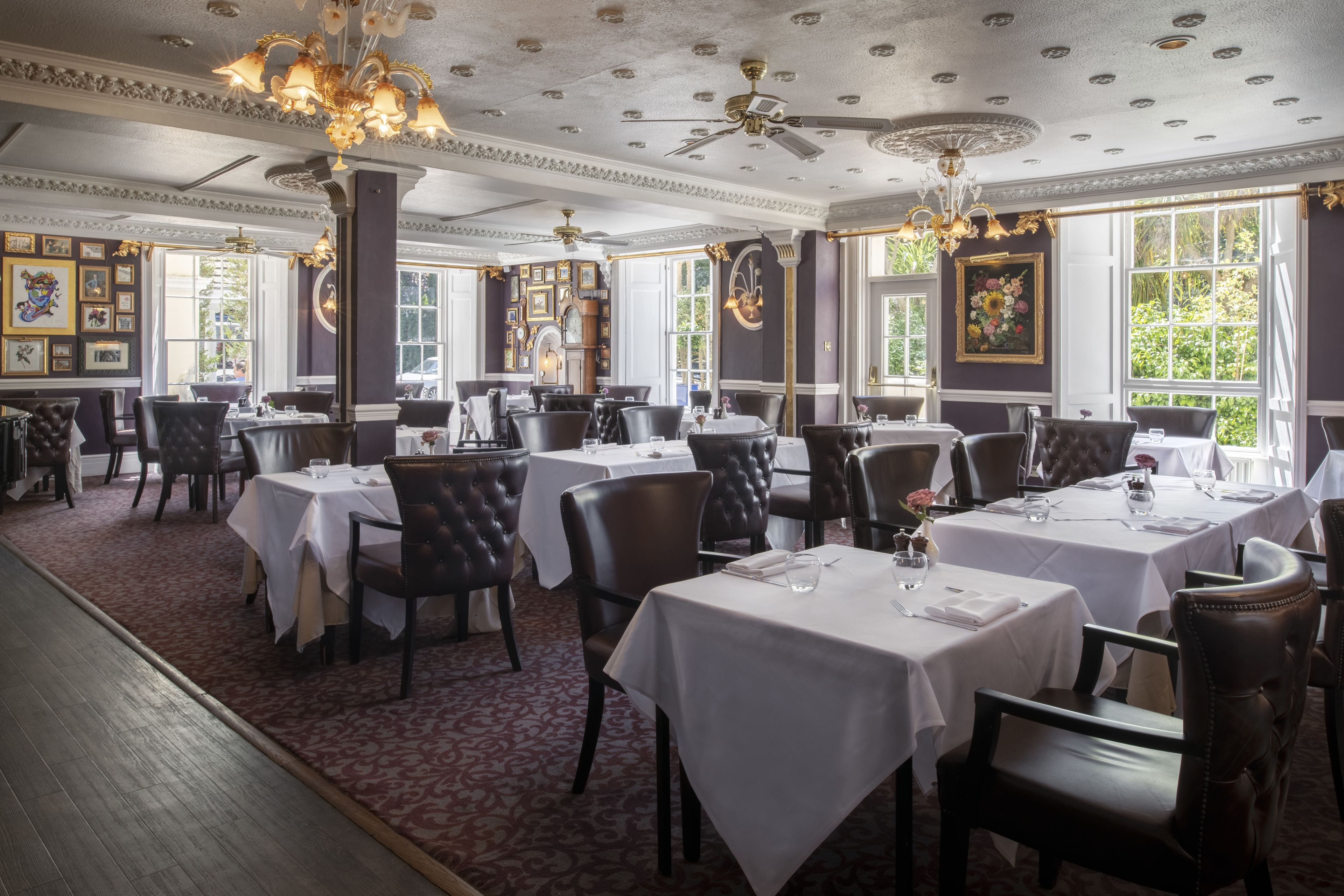 A formal dining room with multiple tables and chairs, ornate ceiling details, and large windows.