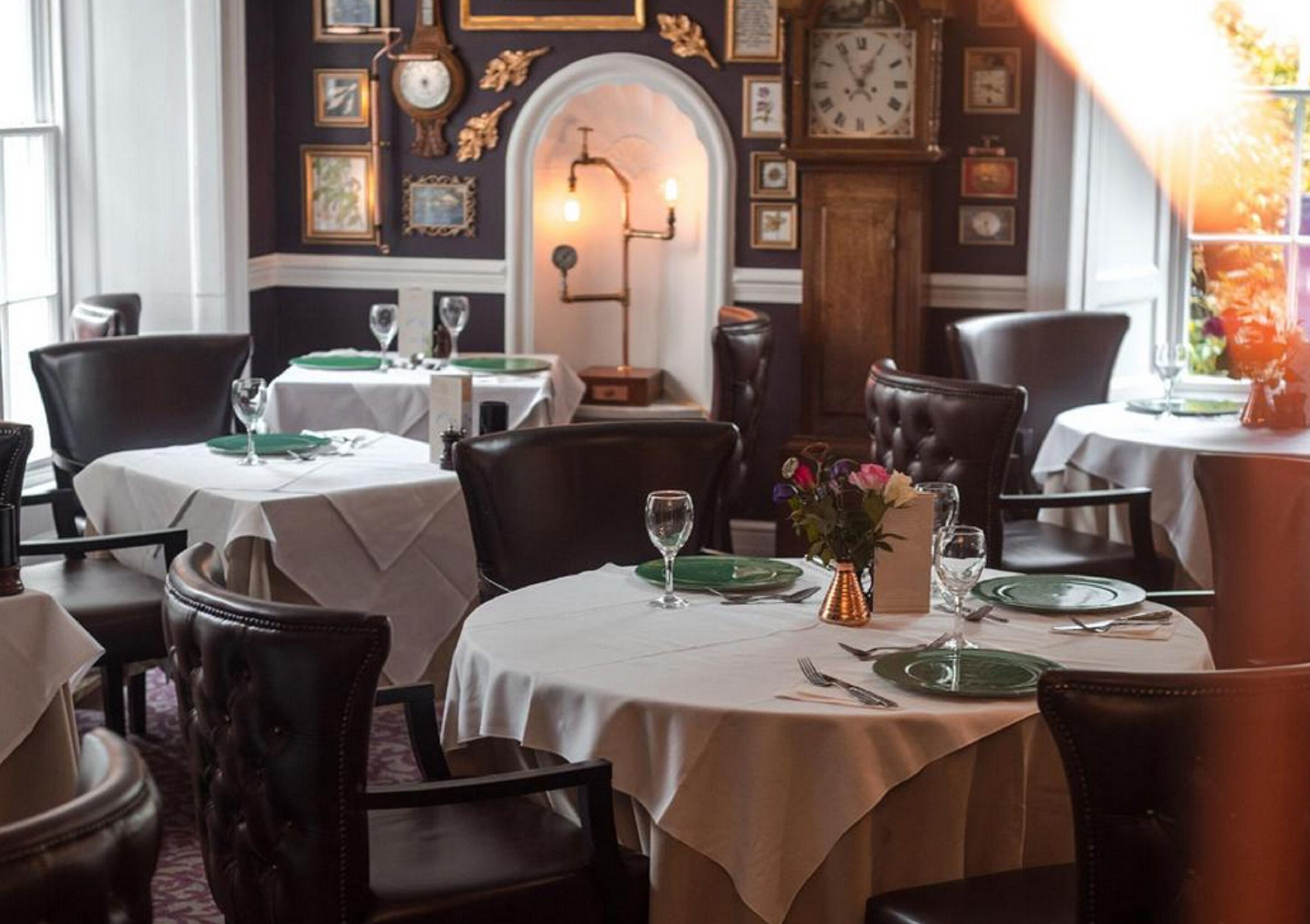 Un comedor con mesas redondas dispuestas para dos, cada una con mantel blanco, platos verdes y cubertería. Alrededor de las mesas hay sillas de cuero. Un jarrón con flores está sobre una de las mesas. Las paredes están decoradas con cuadros y relojes enmarcados.