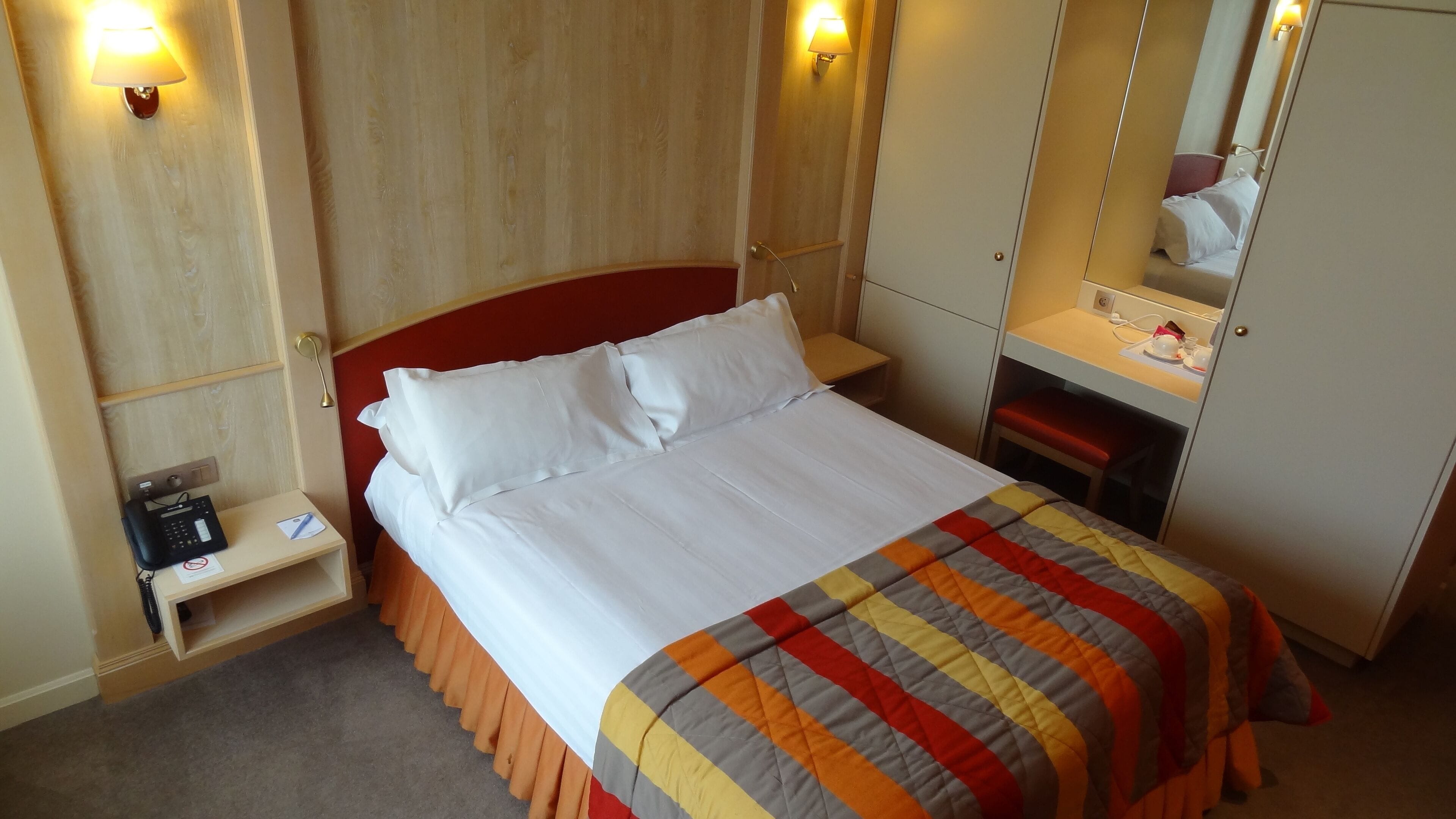 A hotel room with a bed, a bedside table with a phone, a mirror, and a desk with a chair.