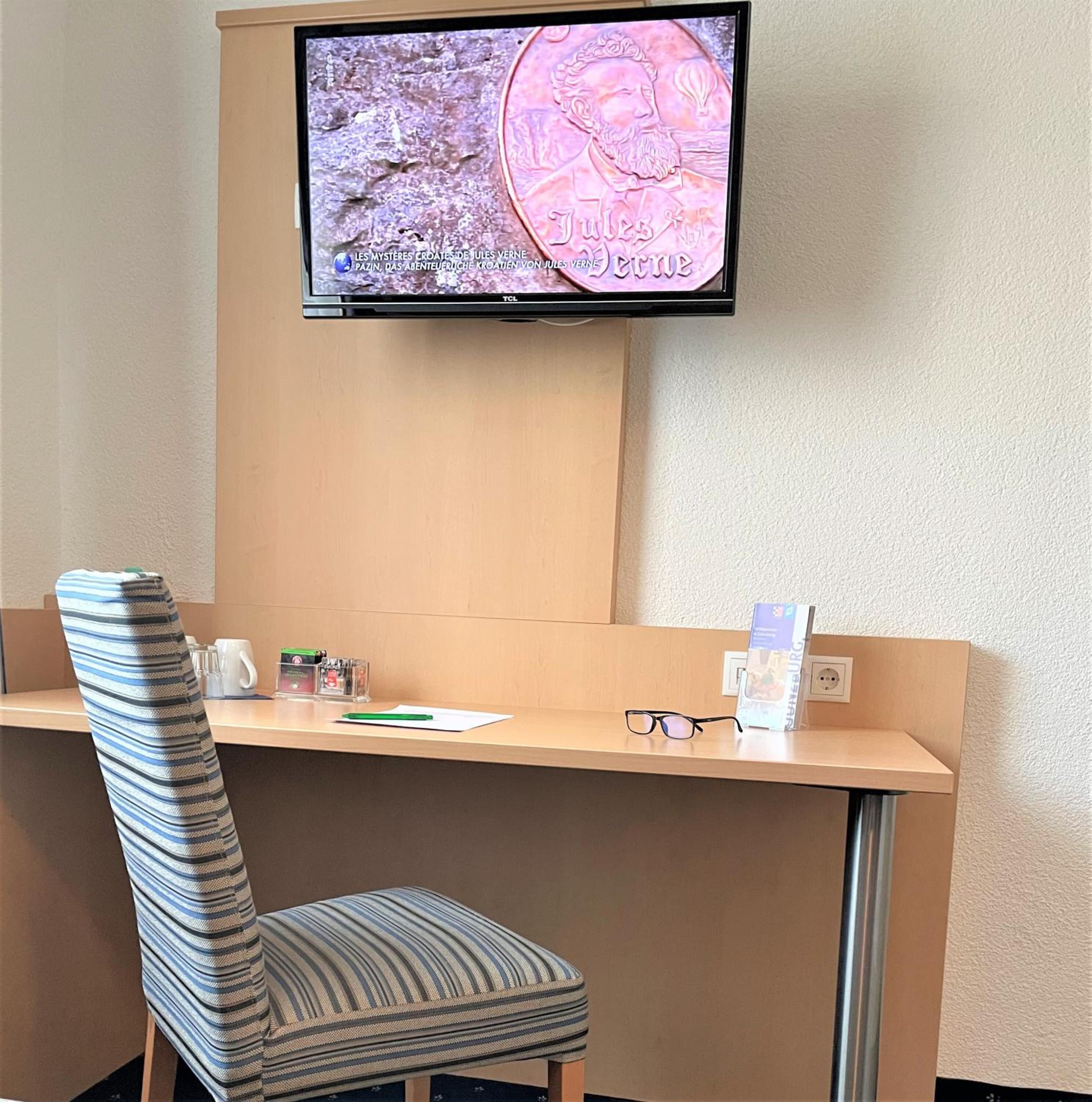 A hotel room with a single desk, a striped chair, a television mounted above the desk, and a few items on the desk including a pen, glasses, and a brochure.