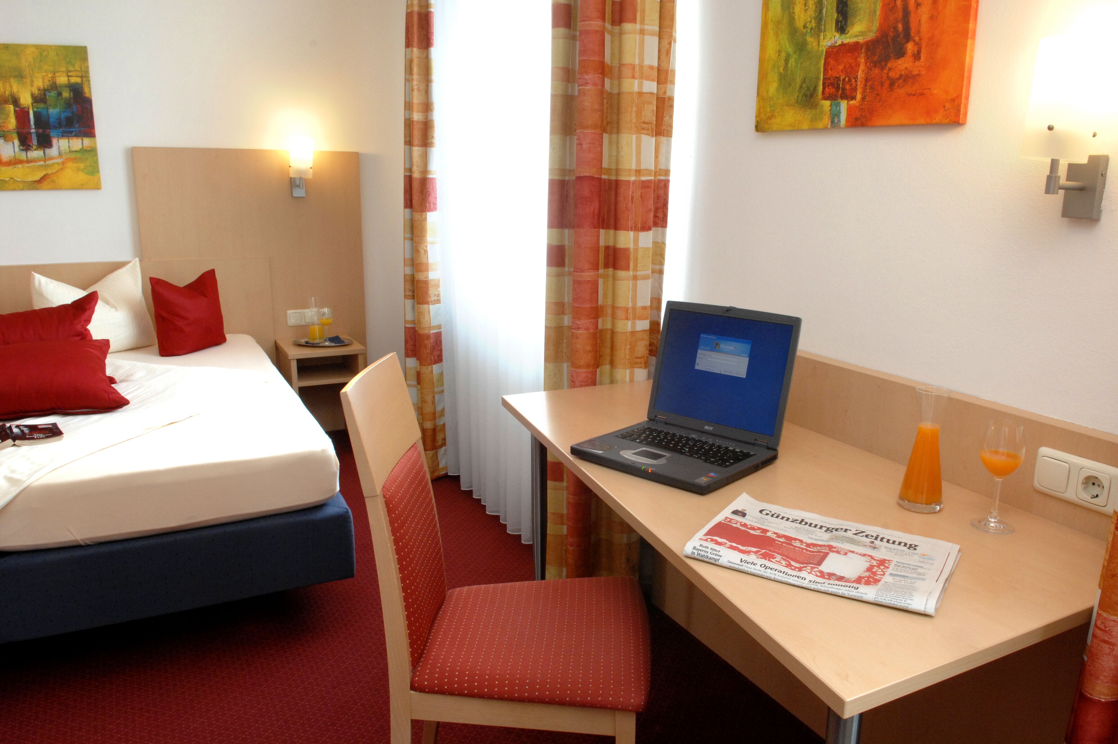 Ein Hotelzimmer mit Bett, Schreibtisch mit Laptop, Stuhl, Tisch mit Zeitung und Orangensaftgläser sowie Nachttisch mit Lampe.