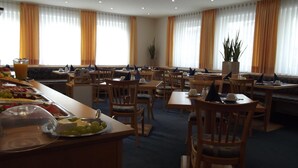 Een restaurantgedeelte in een hotel met verschillende tafels en stoelen, een buffet met fruit en kazen, en grote ramen met gordijnen.