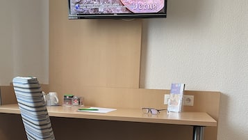 A hotel room with a single desk, a striped chair, a television mounted on the wall, and a small table with a water bottle and glasses.