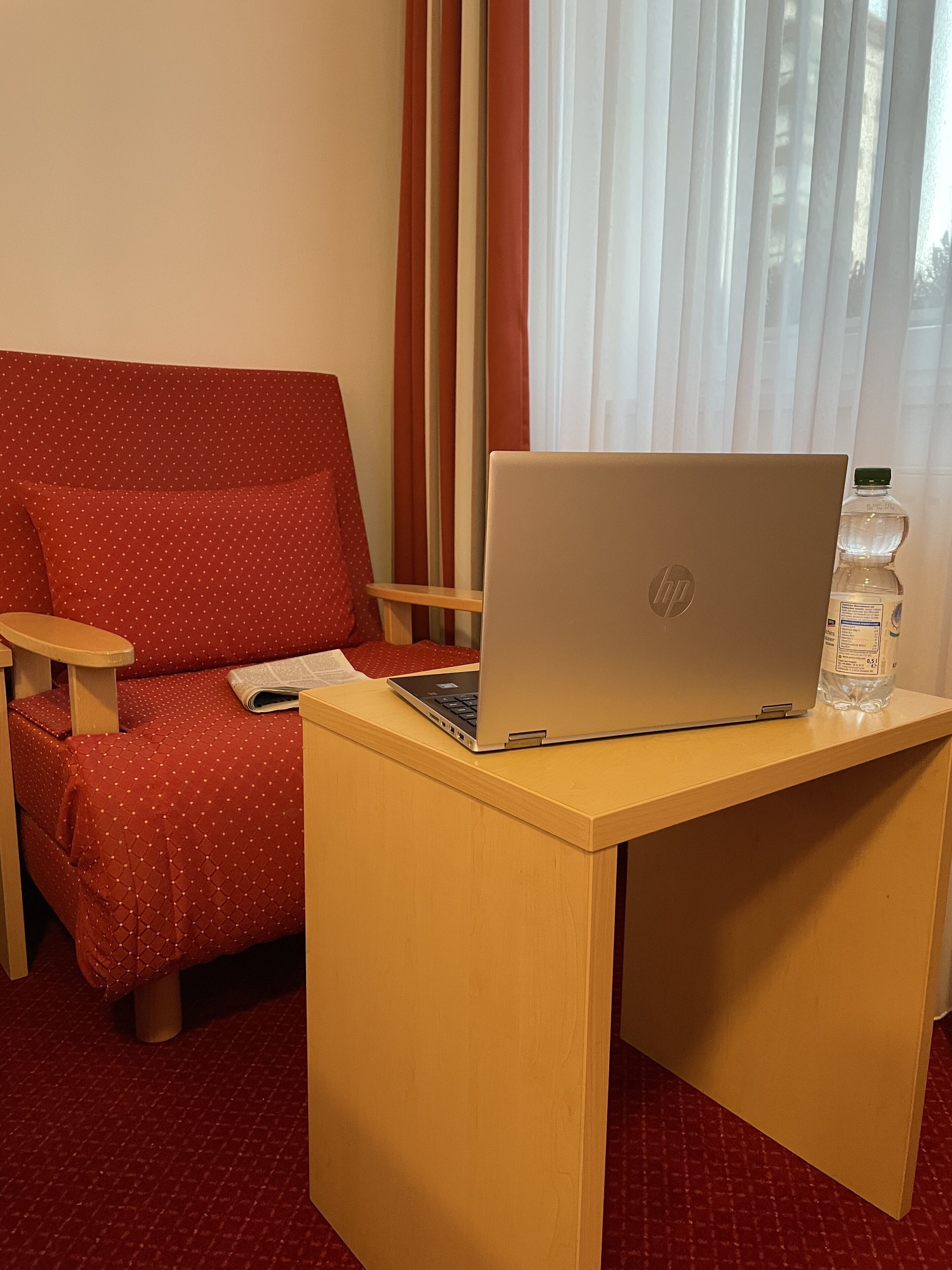 A hotel room with a red polka dot sofa, a wooden desk with a laptop and a water bottle, and a window with sheer curtains.