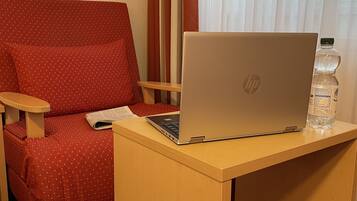 A hotel room with a red polka dot sofa, a wooden desk with a laptop and a water bottle, and a window with sheer curtains.