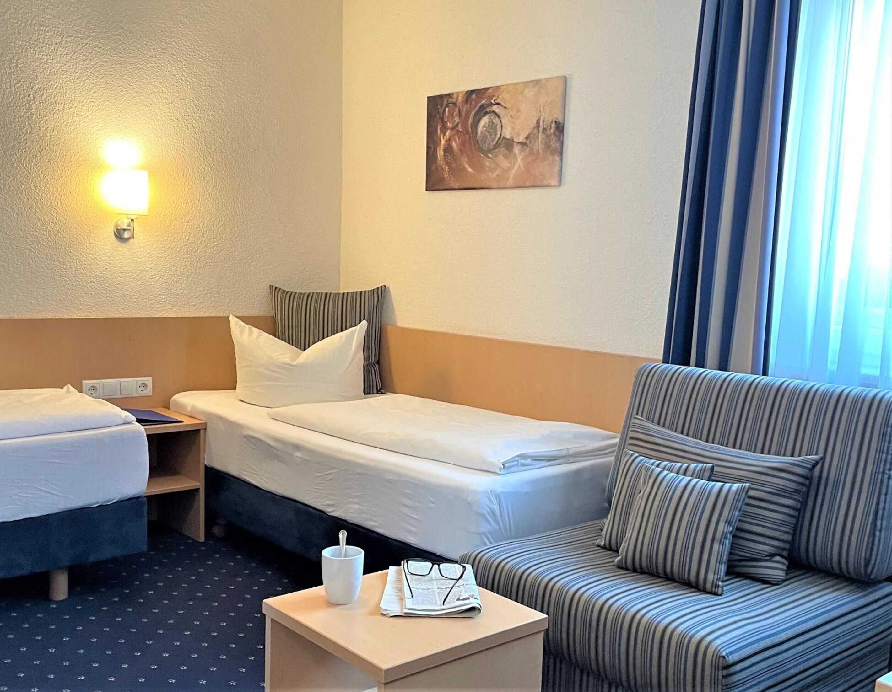 Een hotelkamer met twee bedden, een slaapbank, een klein tafeltje met een kopje en glazen, en een schilderij aan de muur.