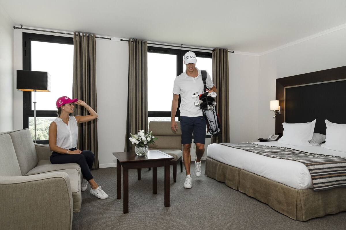 Una habitación de hotel con cama, sofá, una mesita con un florero y una ventana con cortinas.