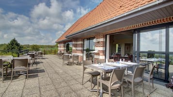Een terras buiten met tafels en stoelen, een bakstenen gebouw met grote ramen en uitzicht op het landschap.