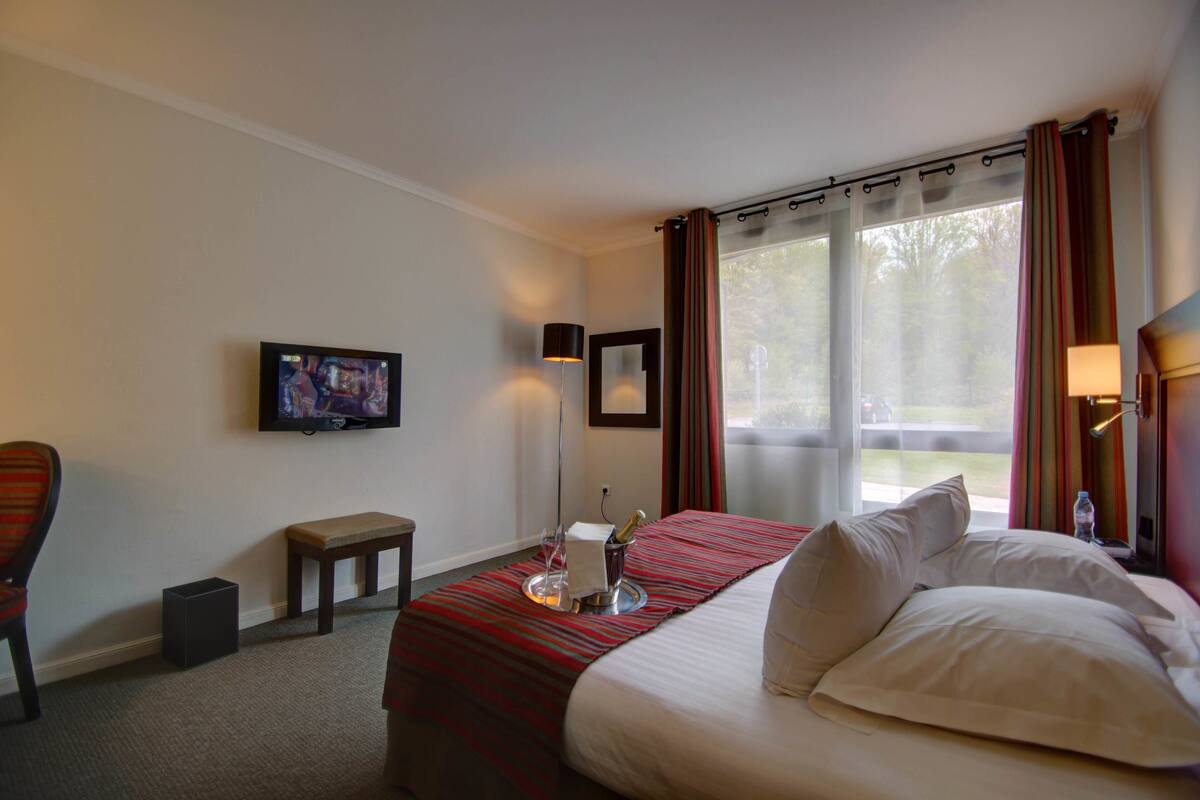 Habitación de hotel con cama, televisión, una mesita con una botella y vasos, y una ventana con cortinas.