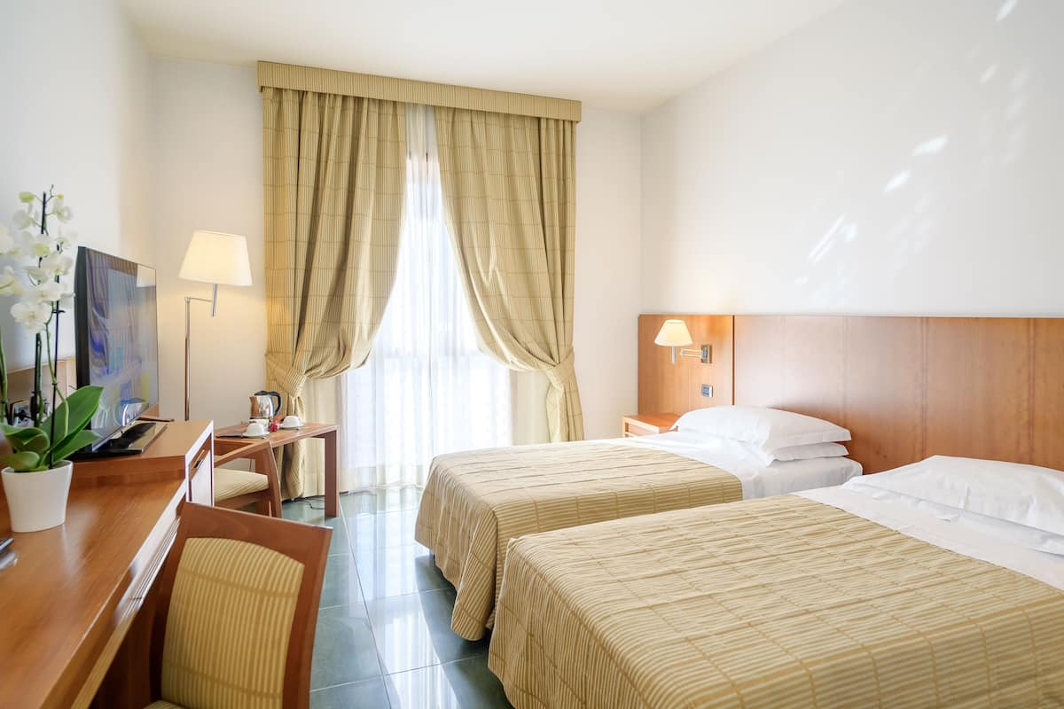 A hotel room with two beds, a desk, a chair, a TV, a lamp, and a window with curtains.