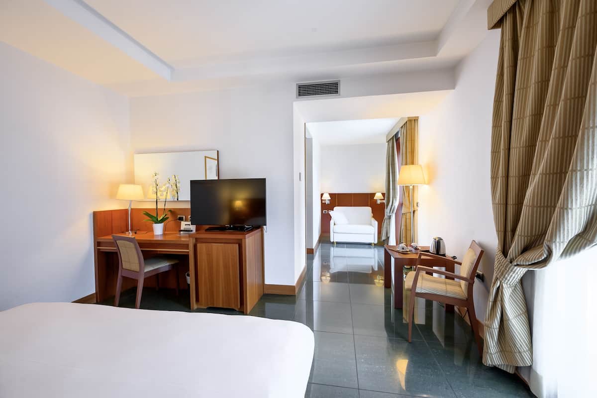 A hotel room with a bed, a desk with a computer, a TV, a chair, a lamp, and a robe hanging on the wall.