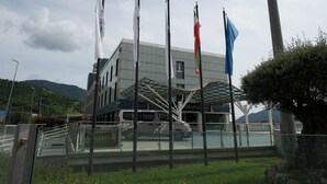 Un bâtiment moderne, orné de plusieurs drapeaux, doté d’une entrée en verre et avec un véhicule garé devant.
