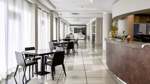 Un hall d’hôtel moderne avec un comptoir d’accueil incurvé, un espace repas avec des tables et des chaises, et de grandes fenêtres dotées de rideaux.
